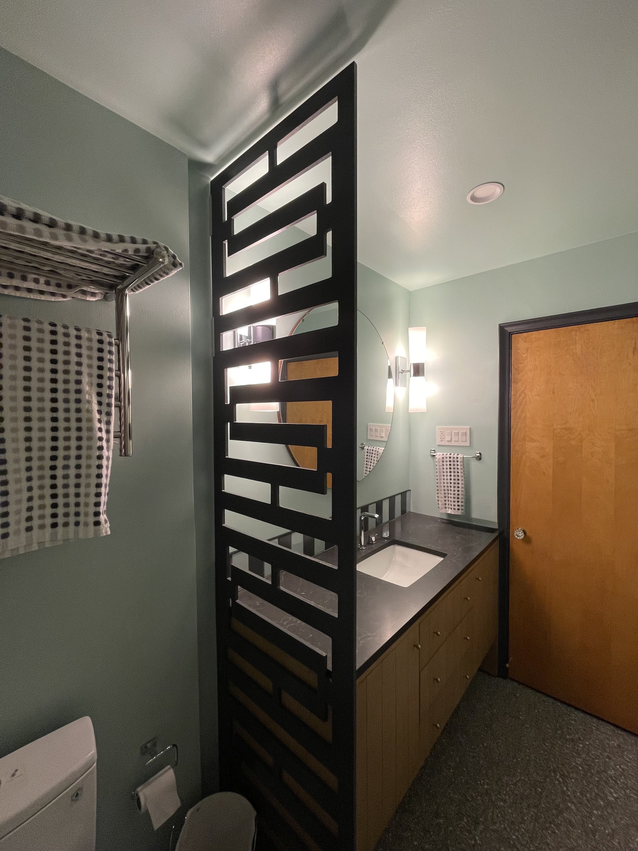 Bathroom with a wooden door, a black decorative room divider, a mirror, a wall light, a black countertop with a white sink, a towel rack with a towel, a light green wall, and a window with a checkered curtain.