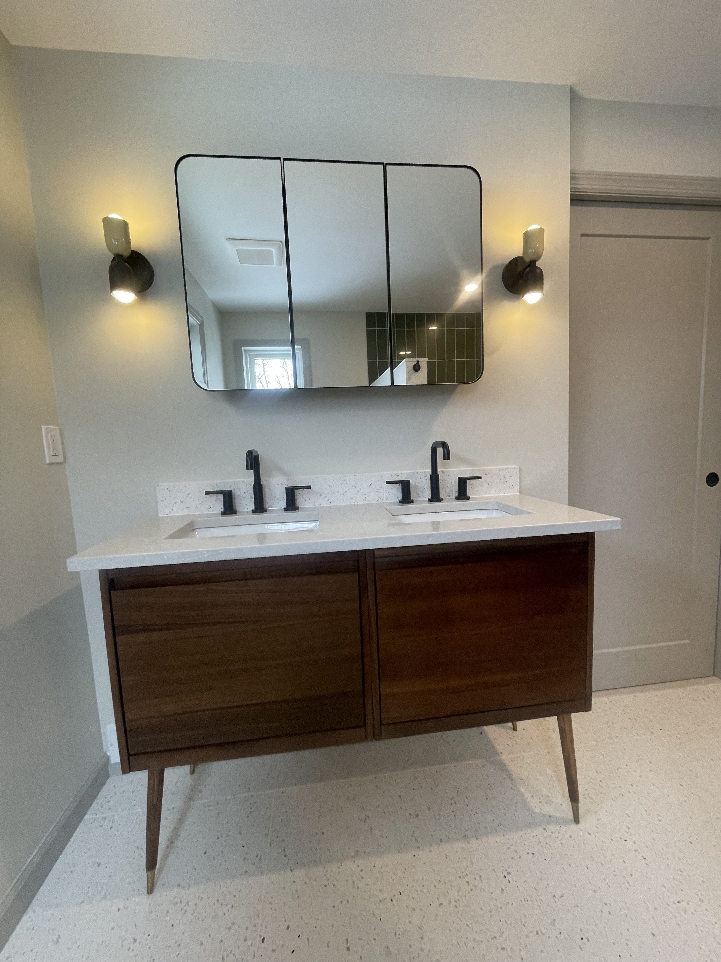 Bathroom with a double sink vanity, black faucets, a large mirror, and green tiled wall in the reflection.