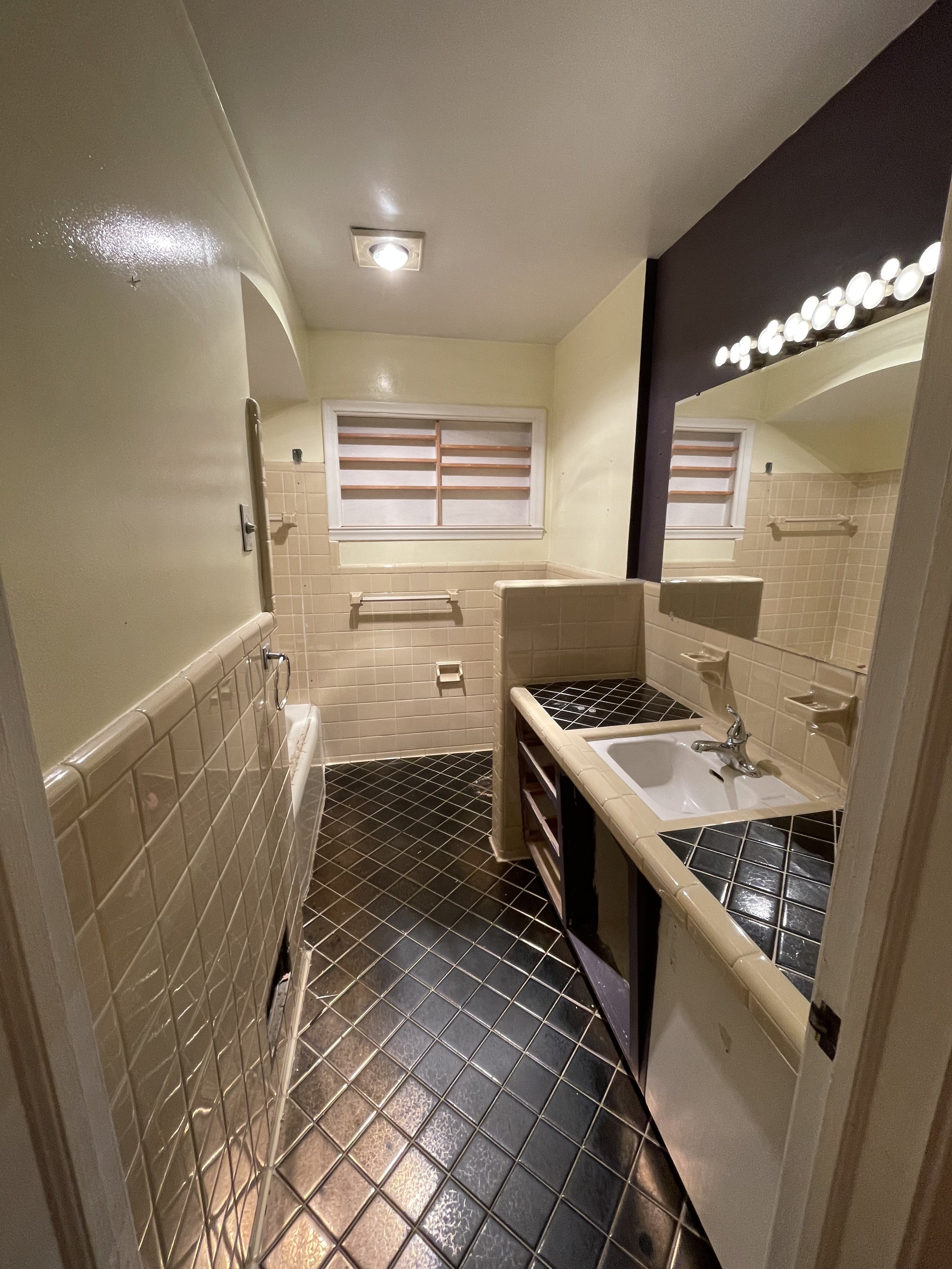 Small bathroom with black tiled floor, yellow tiled counter, small sink, wall mirror with light fixtures, beige tiled walls, and a window with wooden blinds.