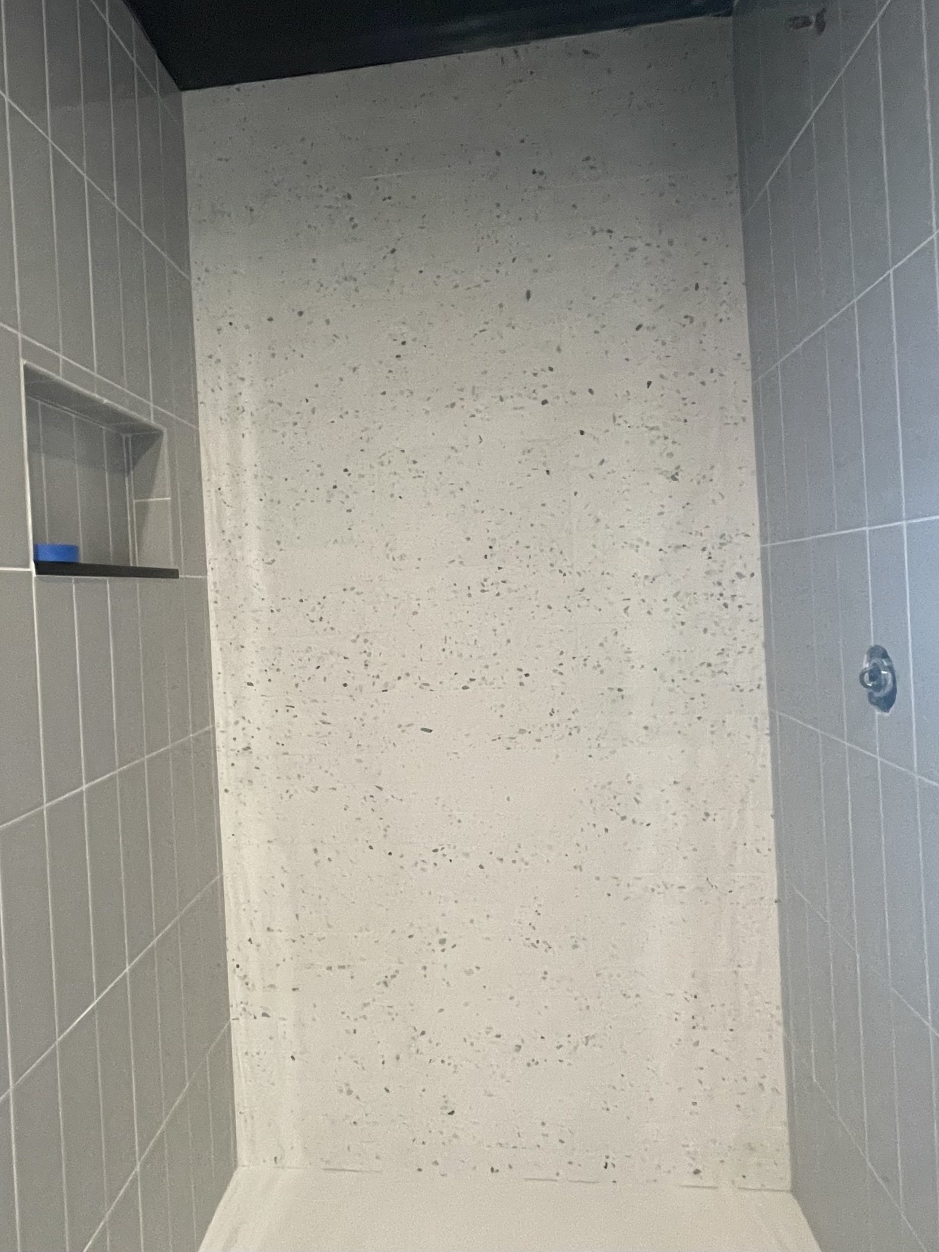 A shower with gray tiles on the left and right walls, a built-in shelf with a blue item inside, a speckled white wall at the back, and a dark ceiling.