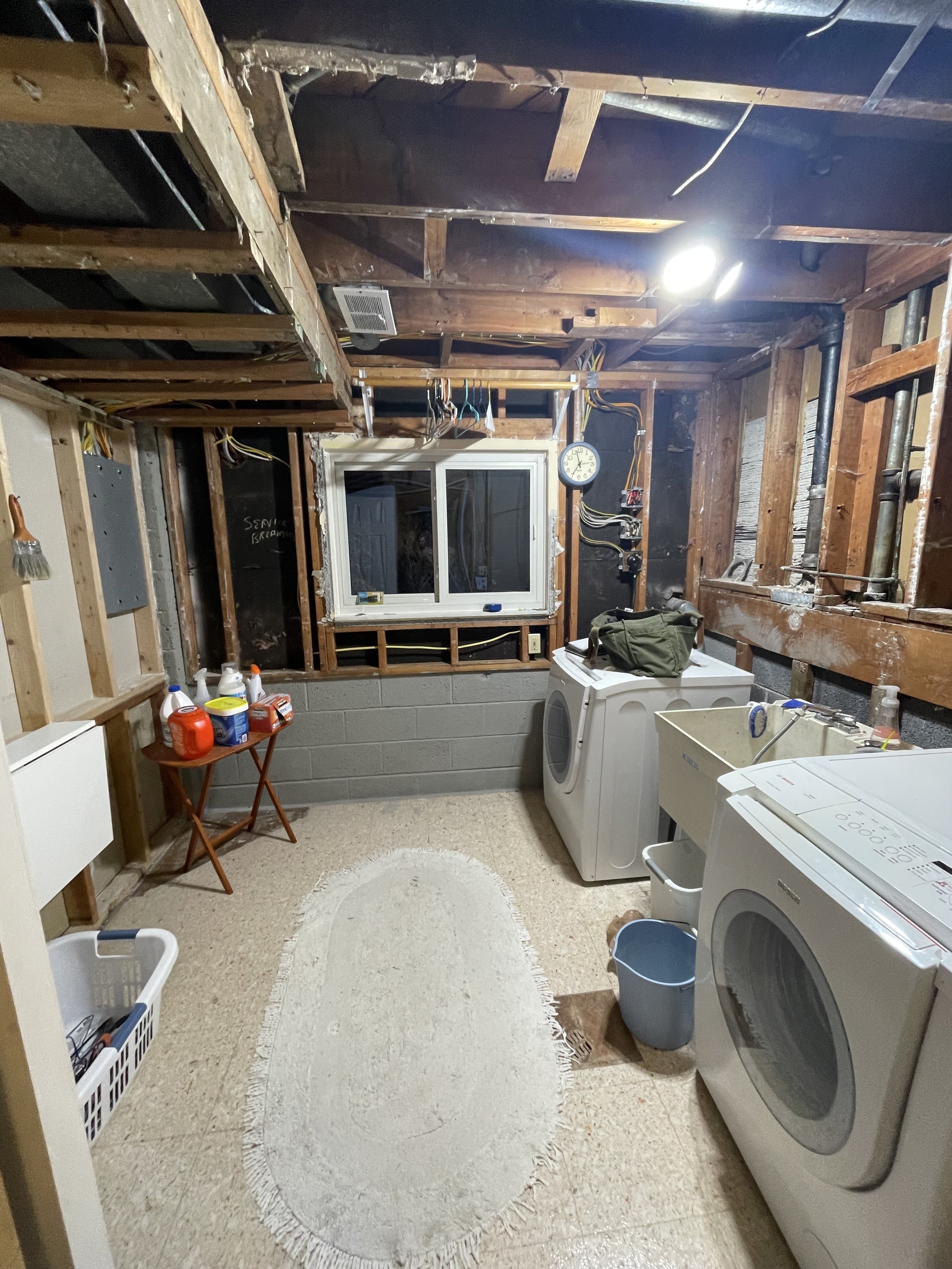 Laundry room in the process of renovation with exposed ceiling beams and wiring, washing machine, dryer, utility sink, window, and cleaning supplies.