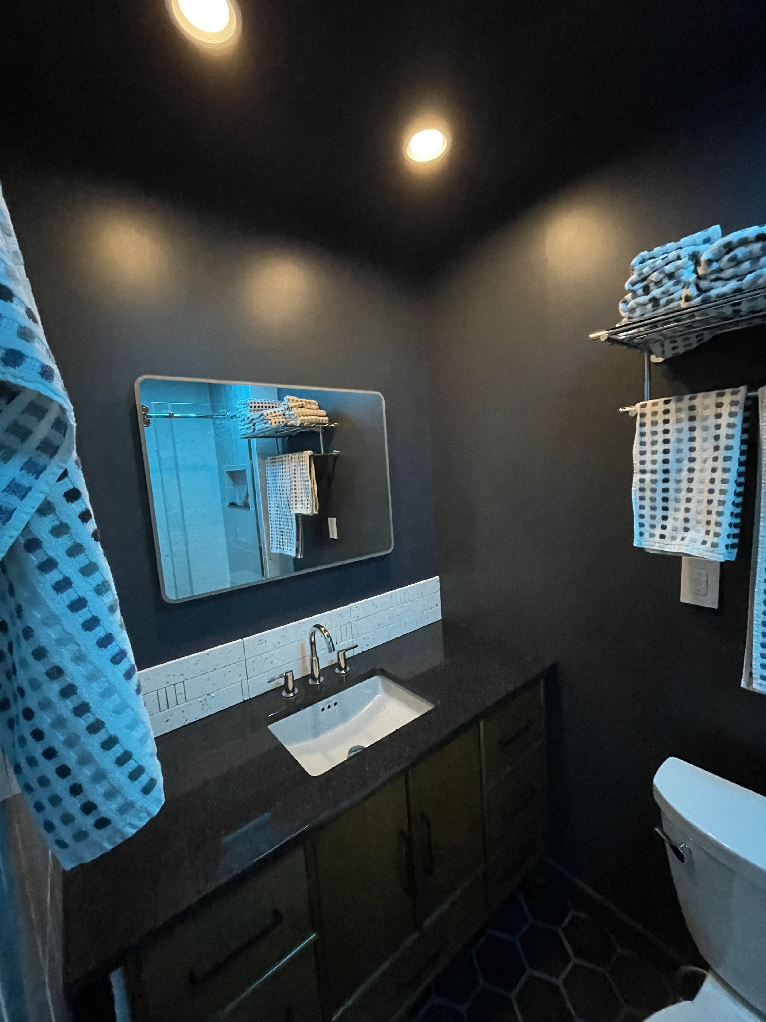 A modern bathroom with dark walls, a white sink and countertop, a mirror, shelves with folded towels, and a toilet.