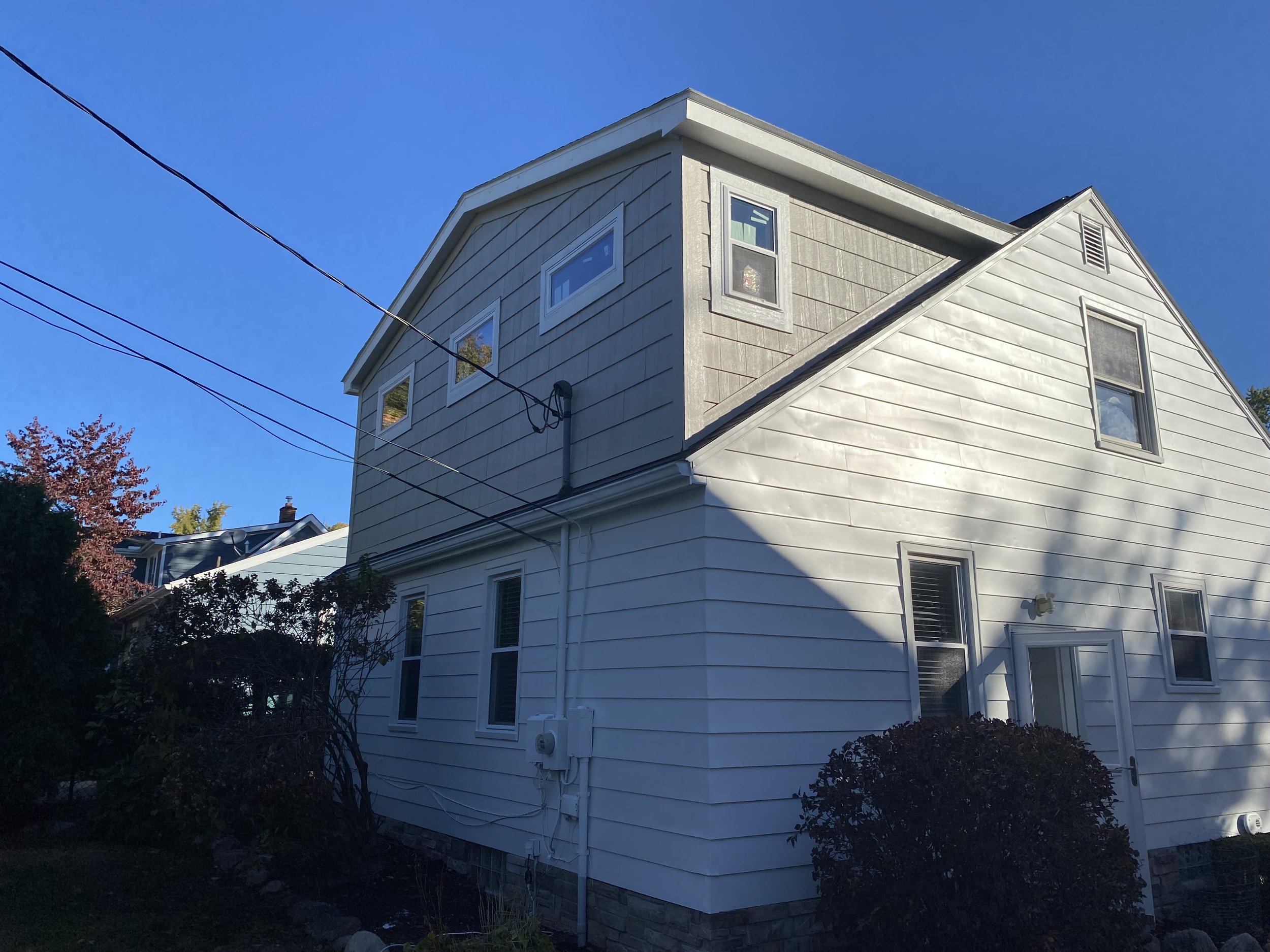 A multi-story house with white and gray siding, multiple windows, and a small shrub in the front yard under a clear blue sky.
