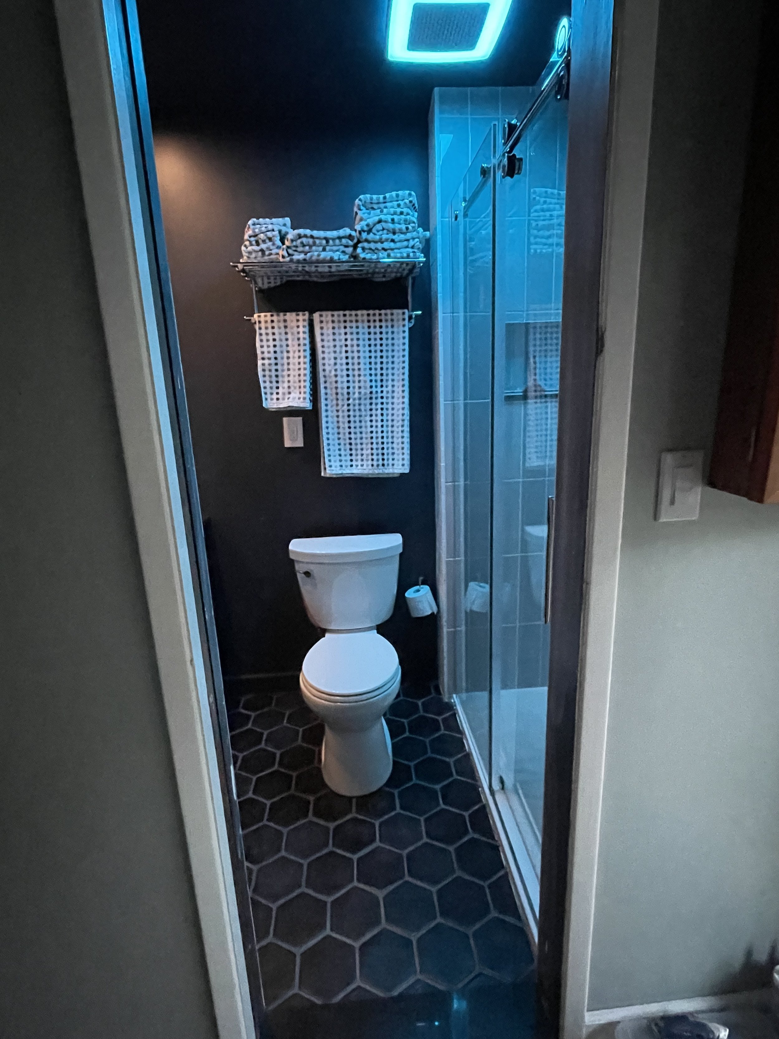 A small bathroom with a white toilet, black hexagonal floor tiles, a shower with a glass door, dark blue walls, two woven baskets with rolled towels on a wall-mounted shelf, two white towels hanging below, and a ceiling vent with blue light.