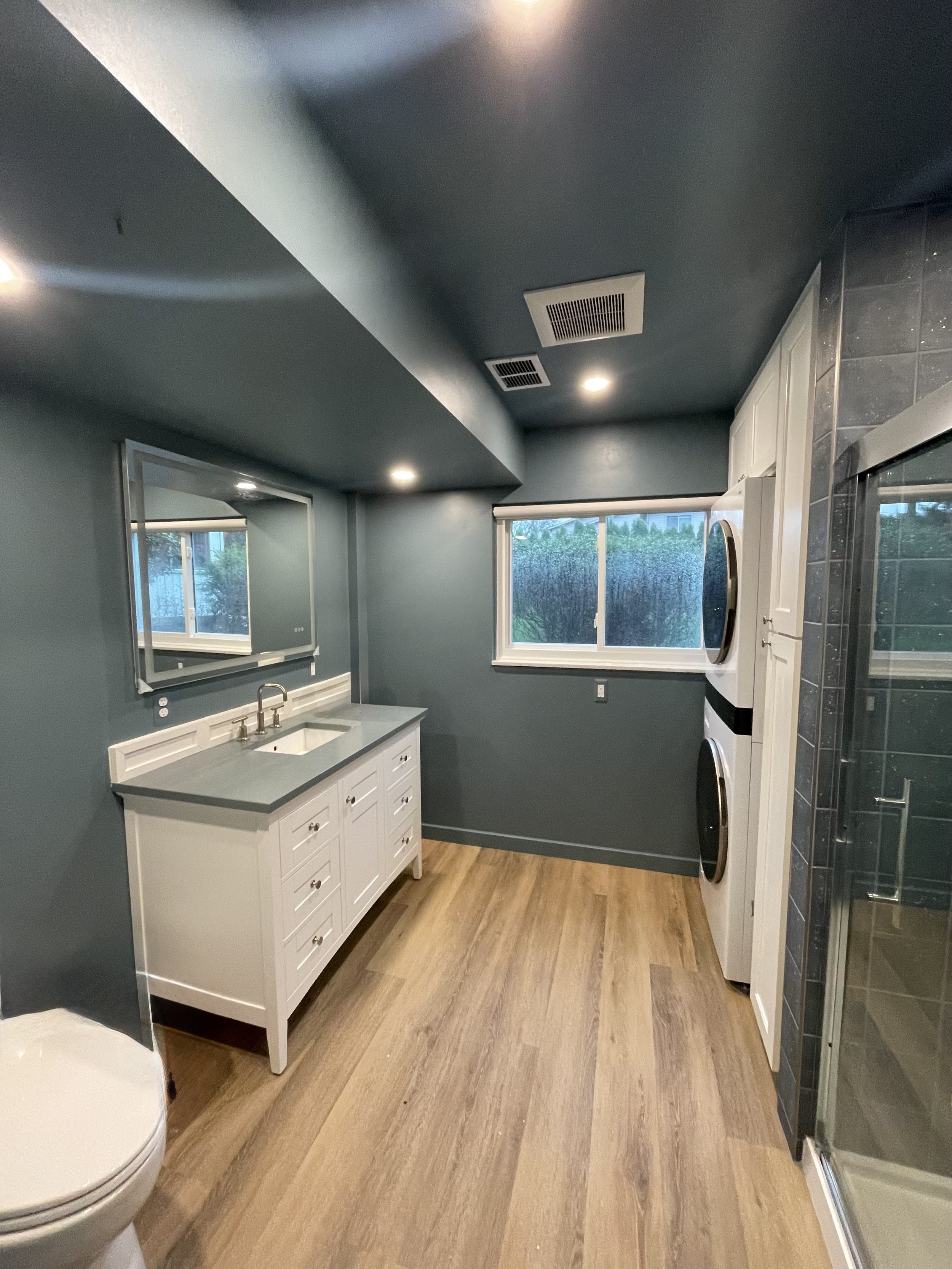 A modern bathroom with gray walls, a white vanity with sink and mirror, a window, a stacked washer and dryer, and a glass shower door.