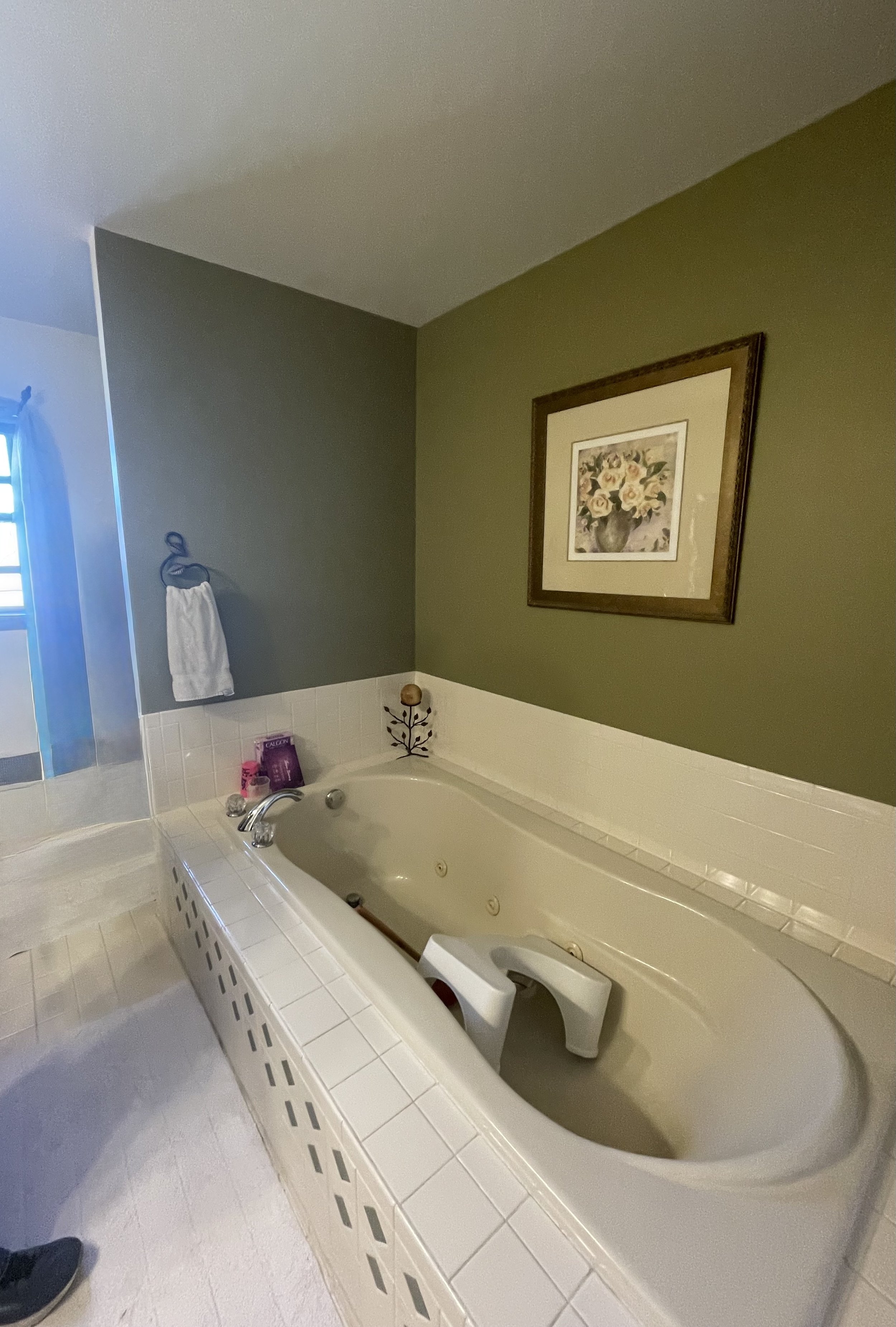 Bathroom with a corner bathtub, green wall, framed floral painting, towel on wall, window with blue curtain.