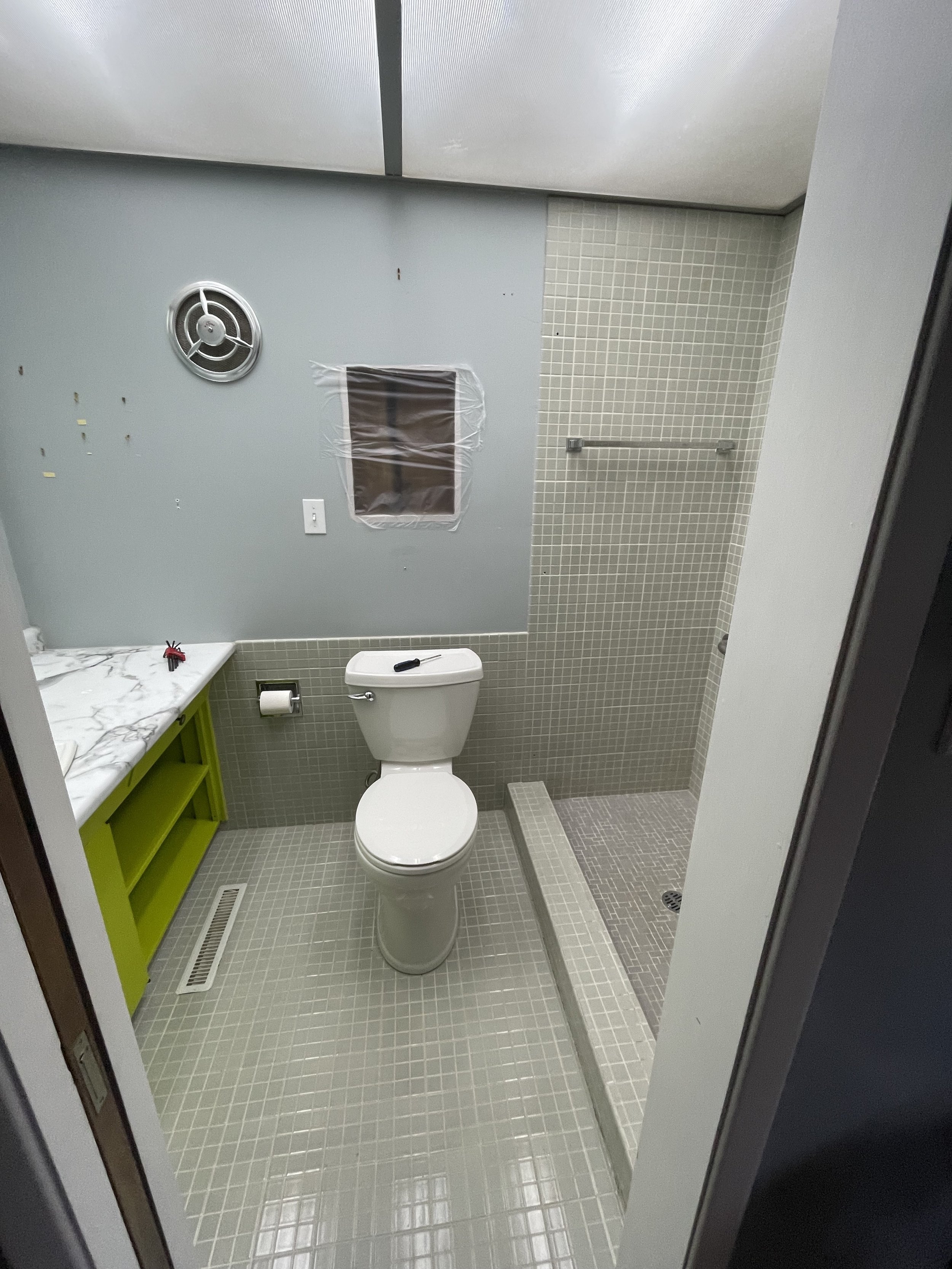 Bathroom with light grey tiled floor, white toilet, green vanity with marble countertop, small window covered with plastic, exhaust fan, towel bar, and a walk-in shower area with shower drain.