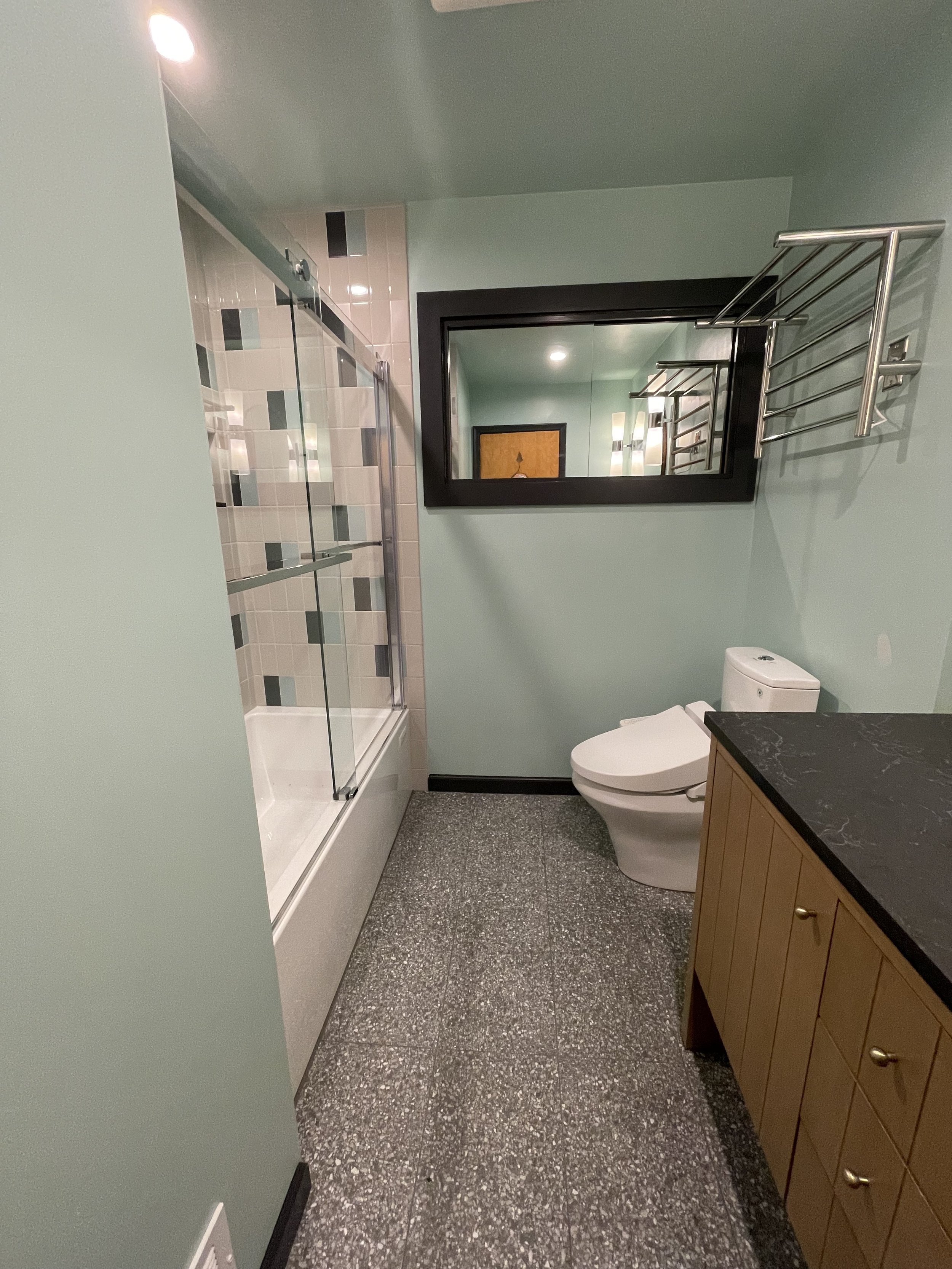 A bathroom with a walk-in shower, toilet, wooden cabinet with a black countertop, a large mirror, and towel racks on the wall, with light green walls and a textured gray floor.