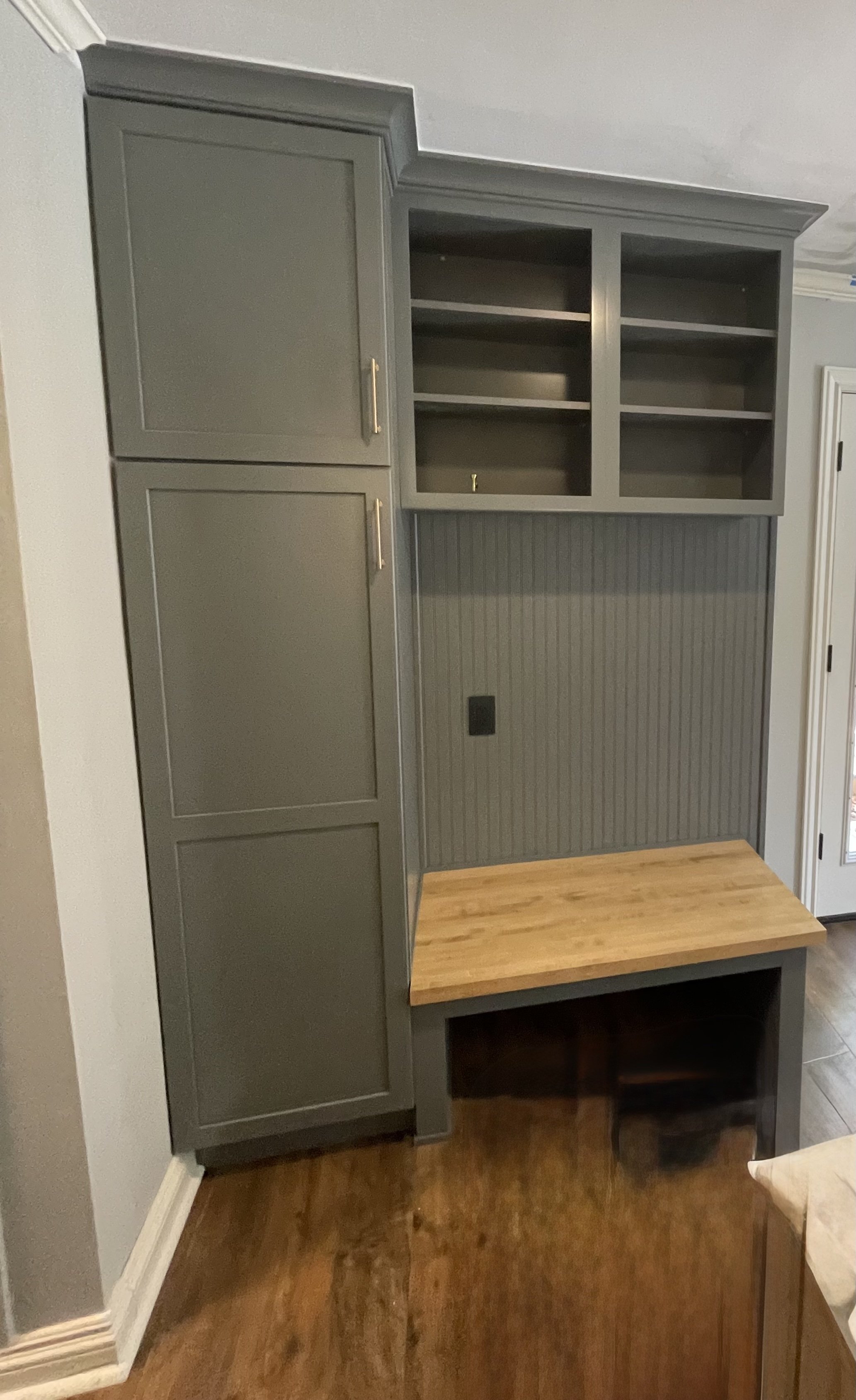 A built-in wall unit with gray cabinets, open shelving, and a wooden bench seat, against a wall with beadboard paneling and a black electrical outlet.