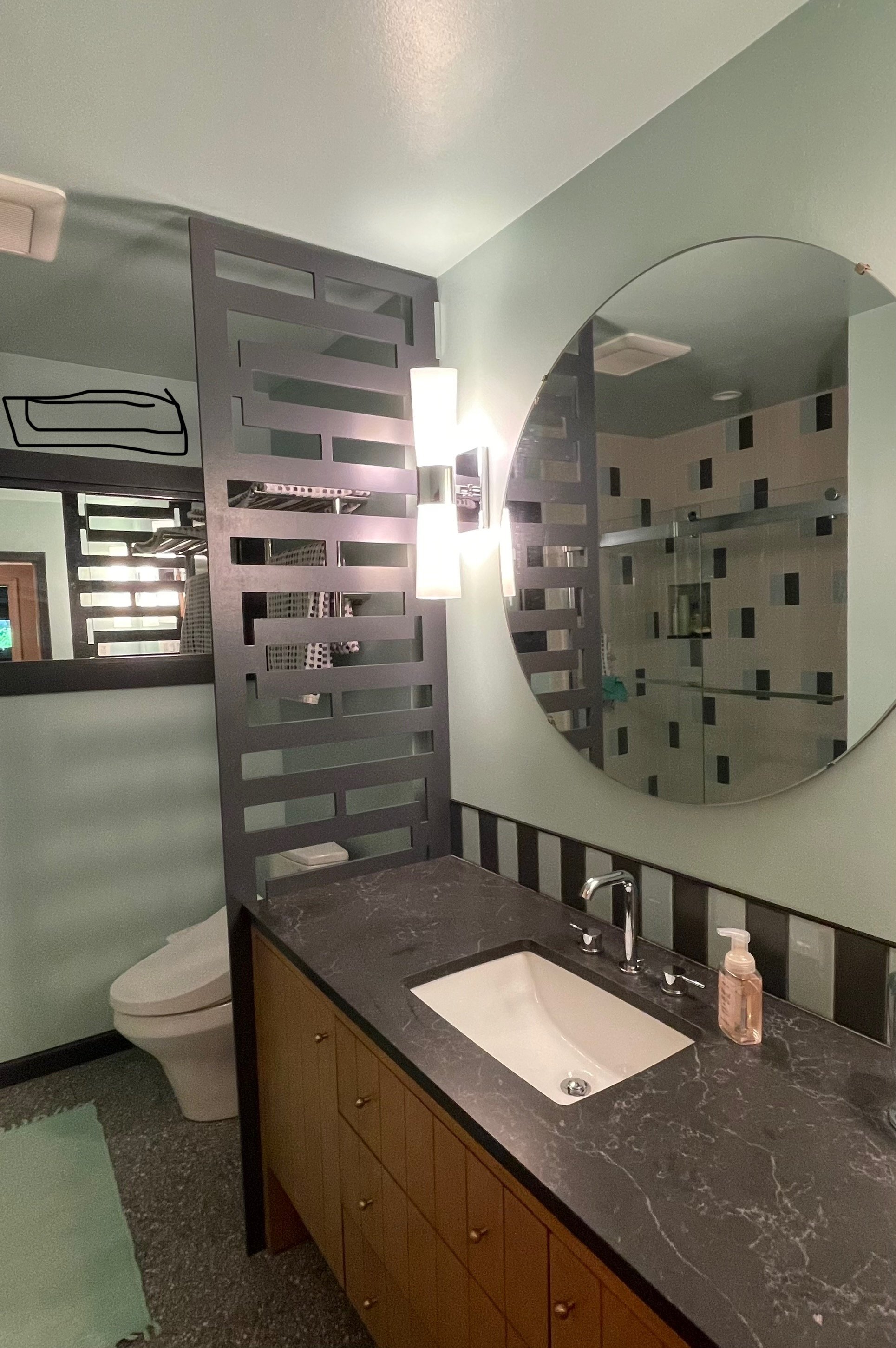 A bathroom with a large round mirror, black and gray textured countertop with a sink, modern wall lamp, a toilet, and a decorative black wood divider.