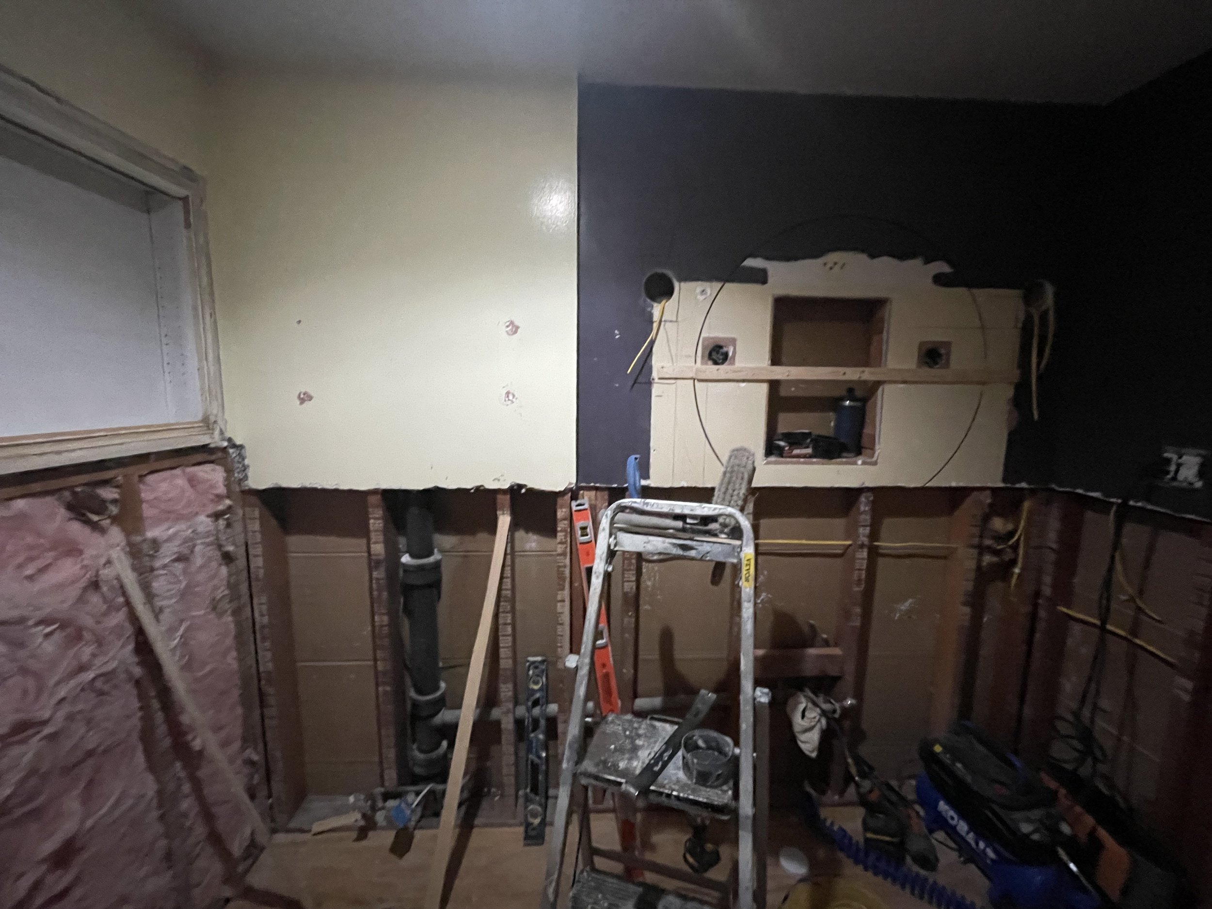 A room under renovation with part of the wall down, showing insulation and pipes. A stepladder and various tools are scattered around.