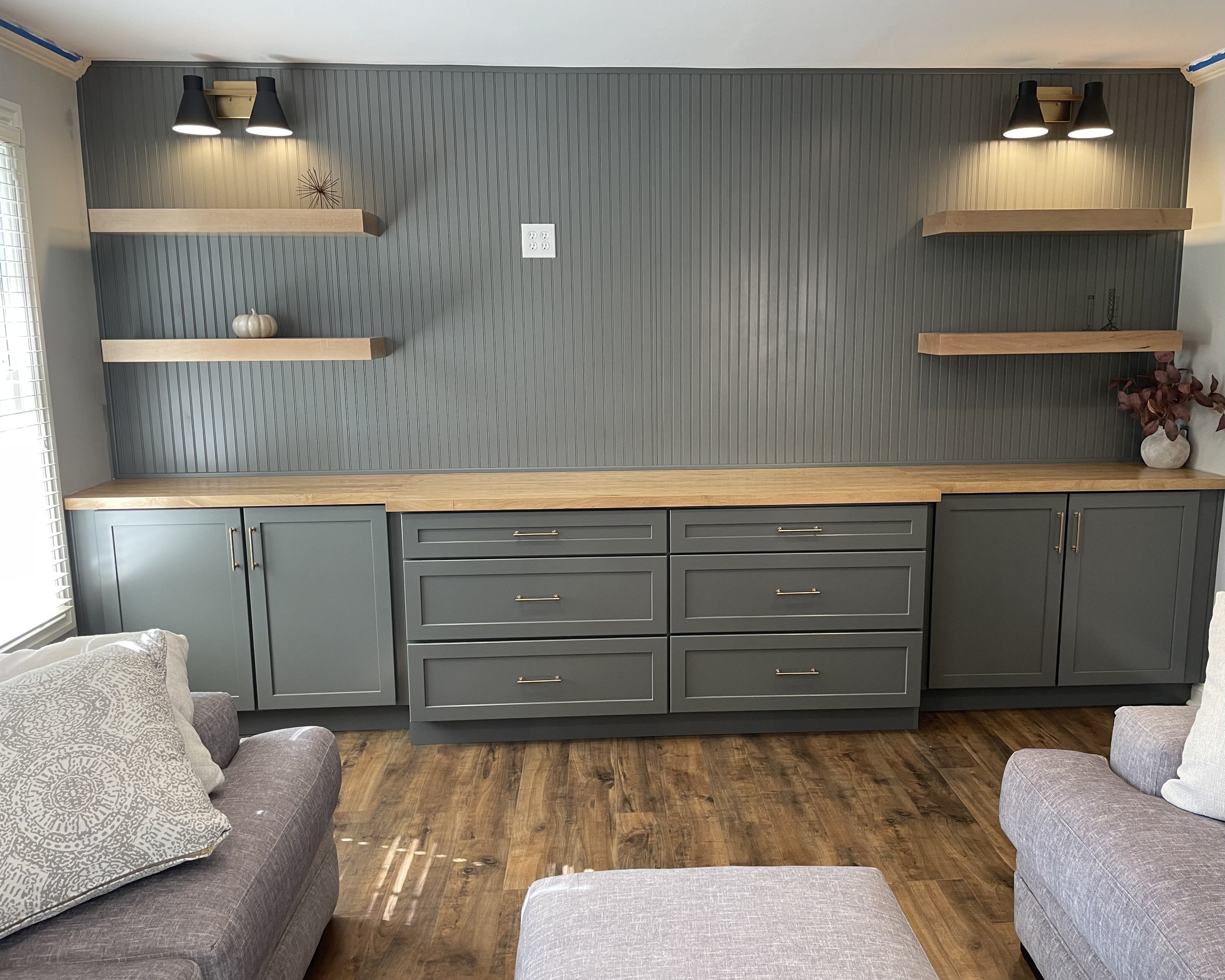 Living room with a built-in cabinet and floating shelves on a blue beadboard wall, with lamps above, a decorative pumpkin, and a vase with flowers on the shelves, and sofas with pillows in the foreground.