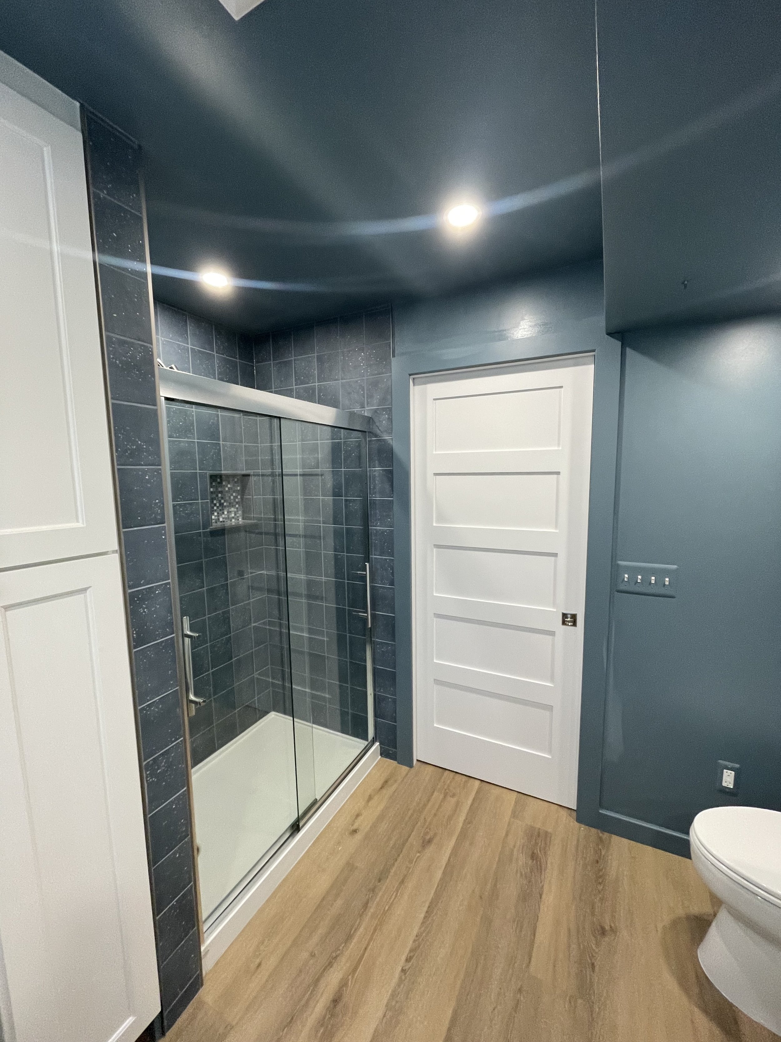 A modern bathroom with blue walls, a glass shower with dark gray tiles, wooden flooring, a white door, and a part of a toilet visible.