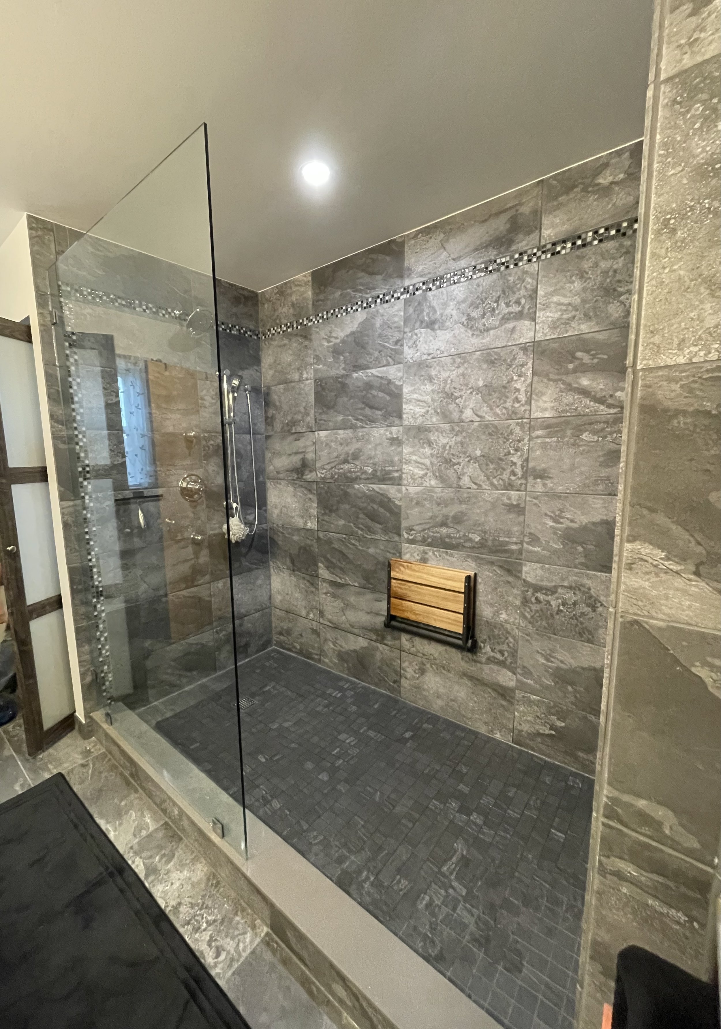 Modern walk-in shower with gray tiled walls and black tile floor, glass enclosure, small built-in wooden bench, and shower fixtures.