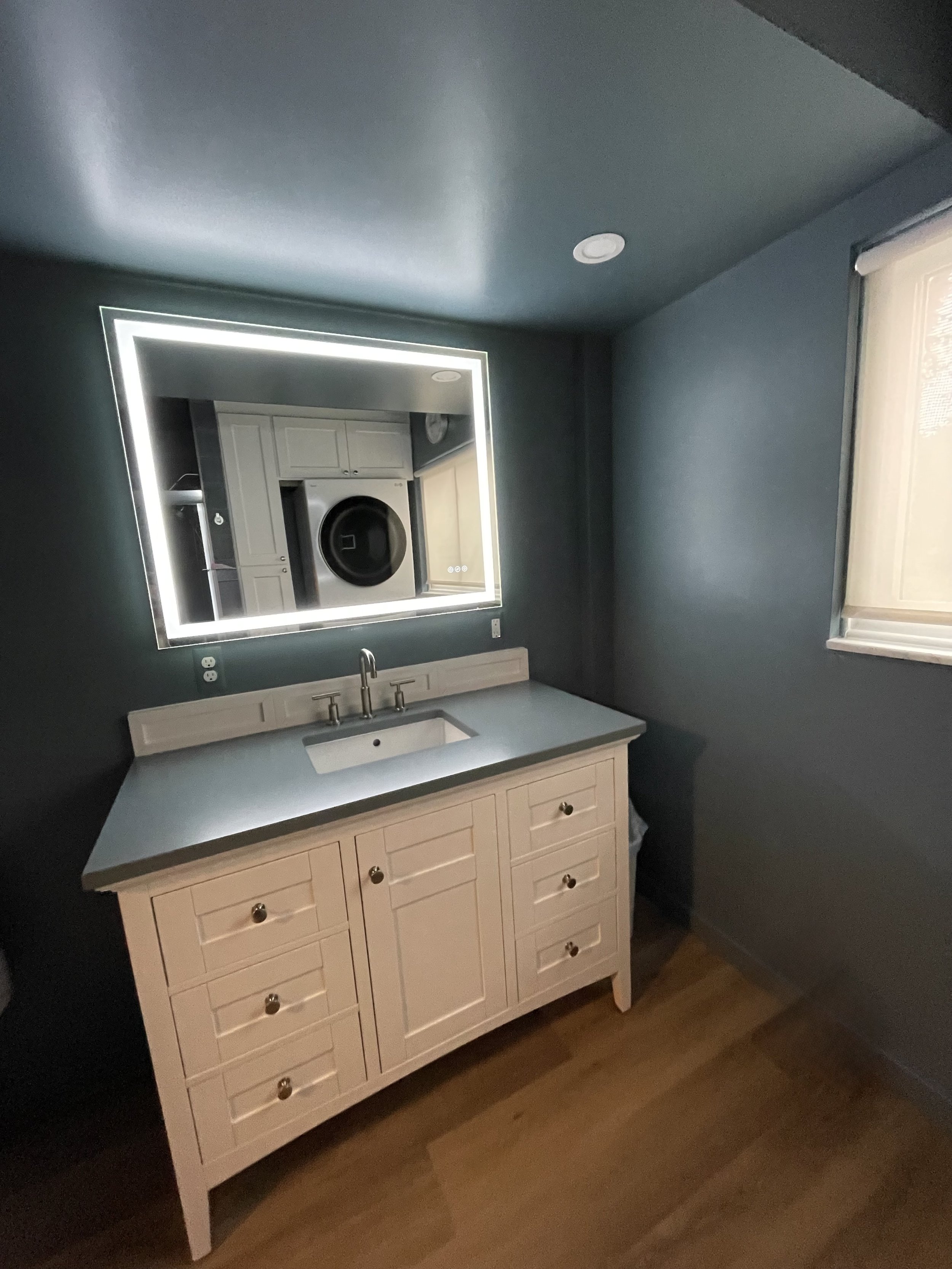A bathroom vanity with a sink and a mirror with LED lighting, reflected in the mirror is a laundry washer inside a room.