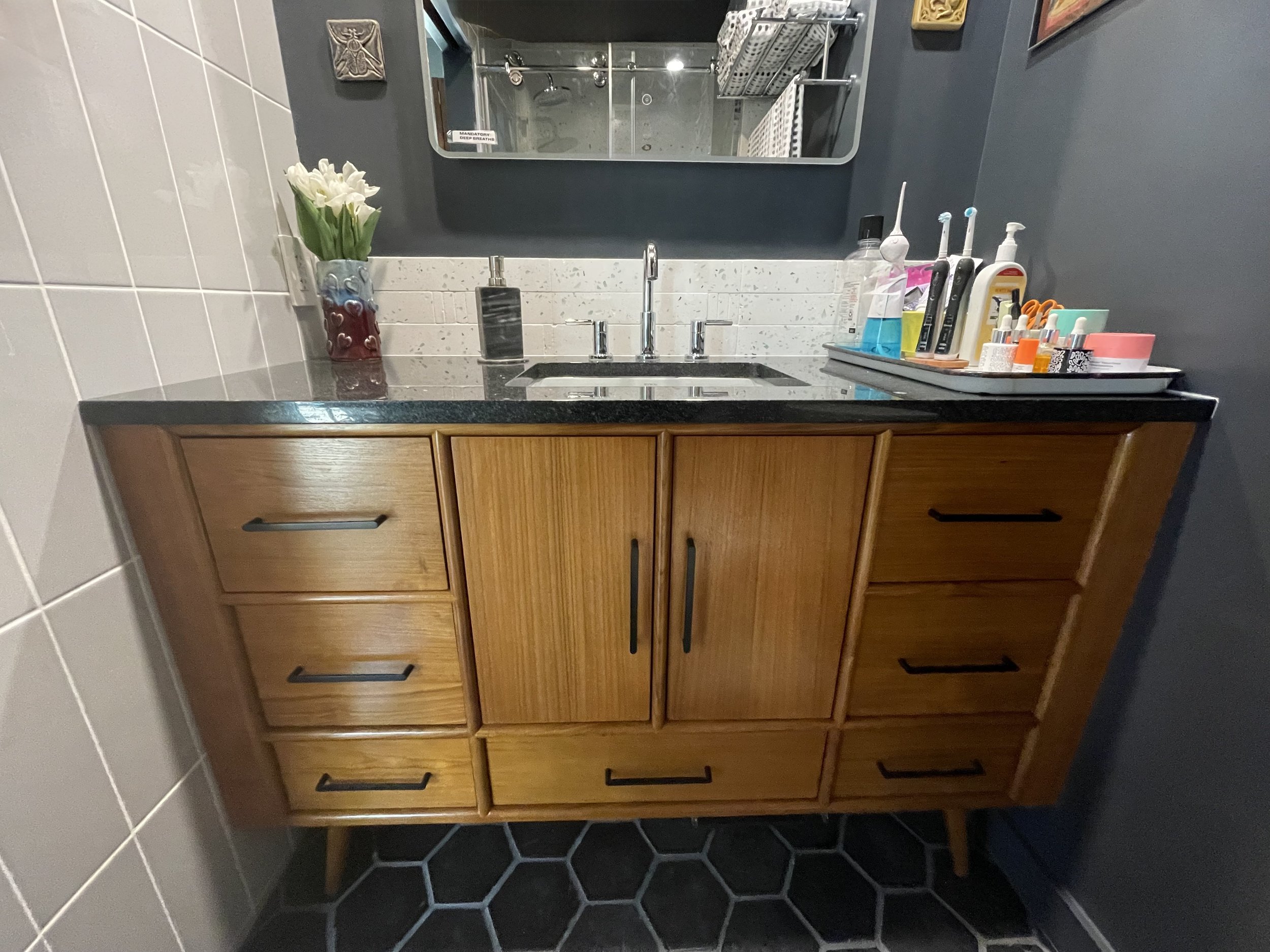A bathroom vanity with a black countertop, a rectangular sink, and various toiletries, against a dark wall with a mirror above.