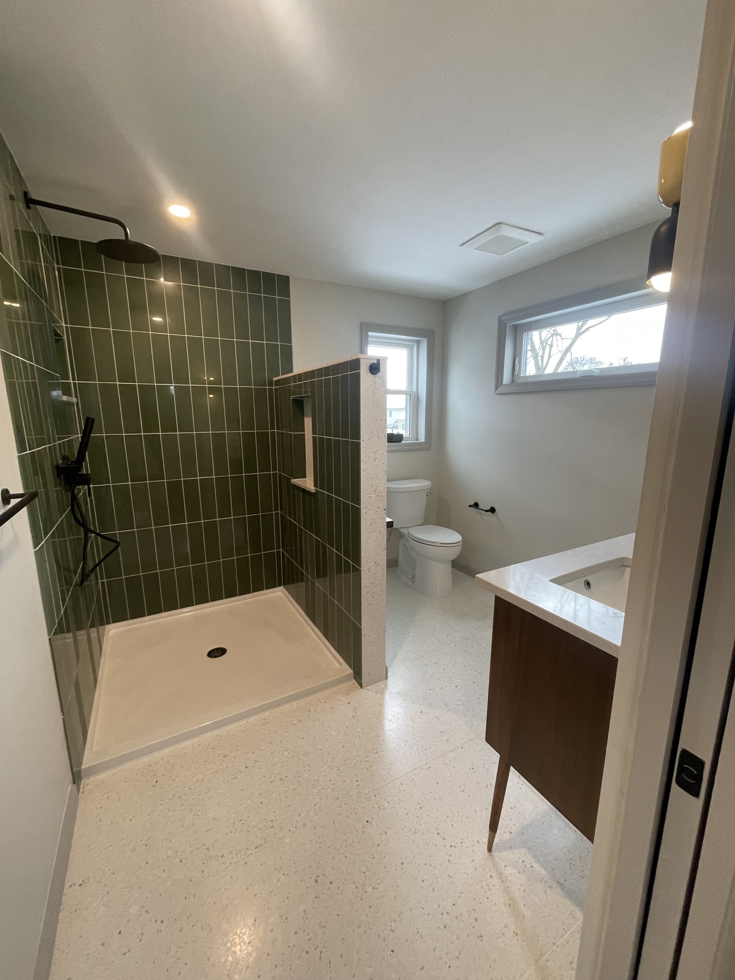 A modern bathroom with a walk-in shower with dark green tiles, a white toilet, a small window, and a wooden vanity with a sink.