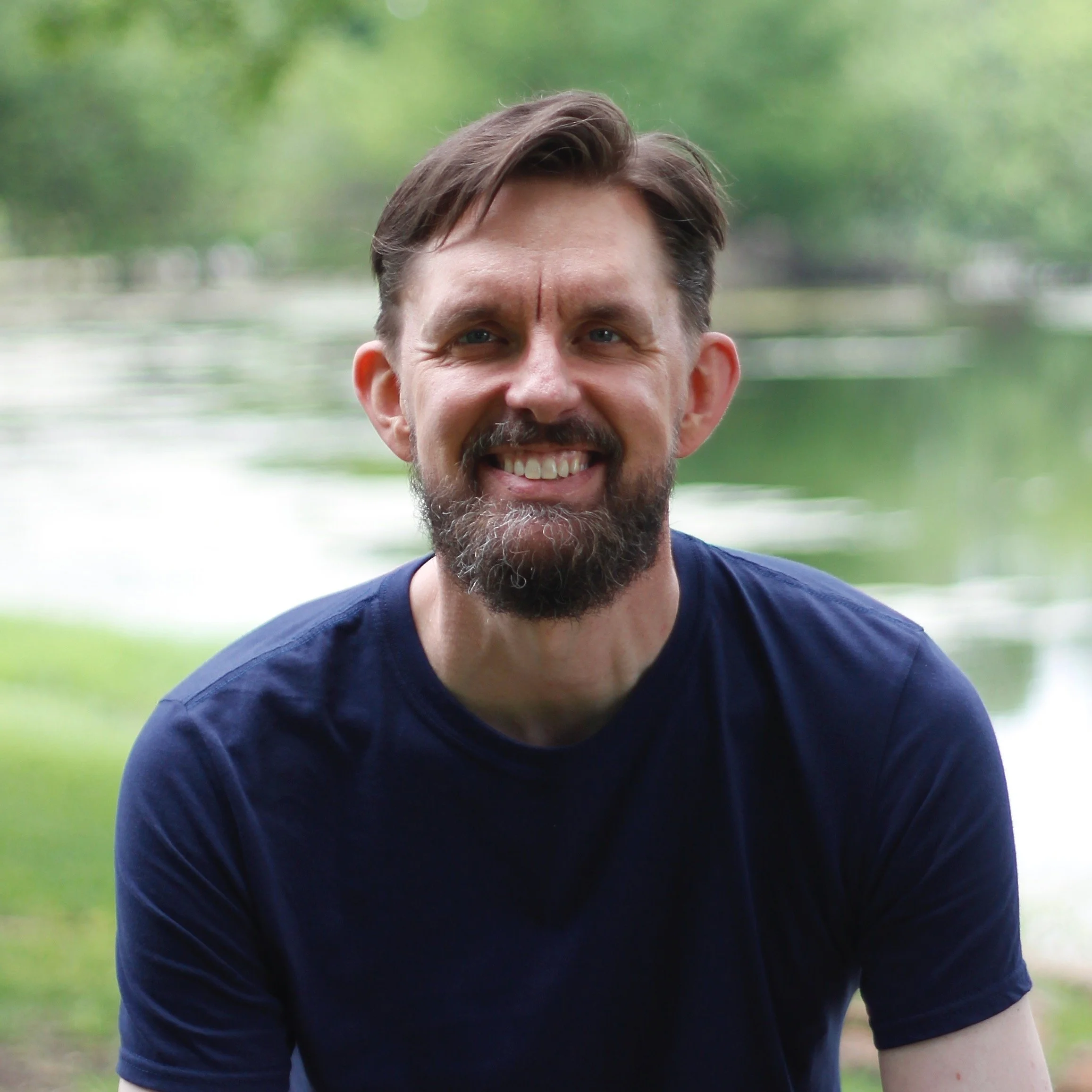 A smiling man with a beard and mustache, wearing a navy blue t-shirt, outdoors near a lake with green trees in the background.
