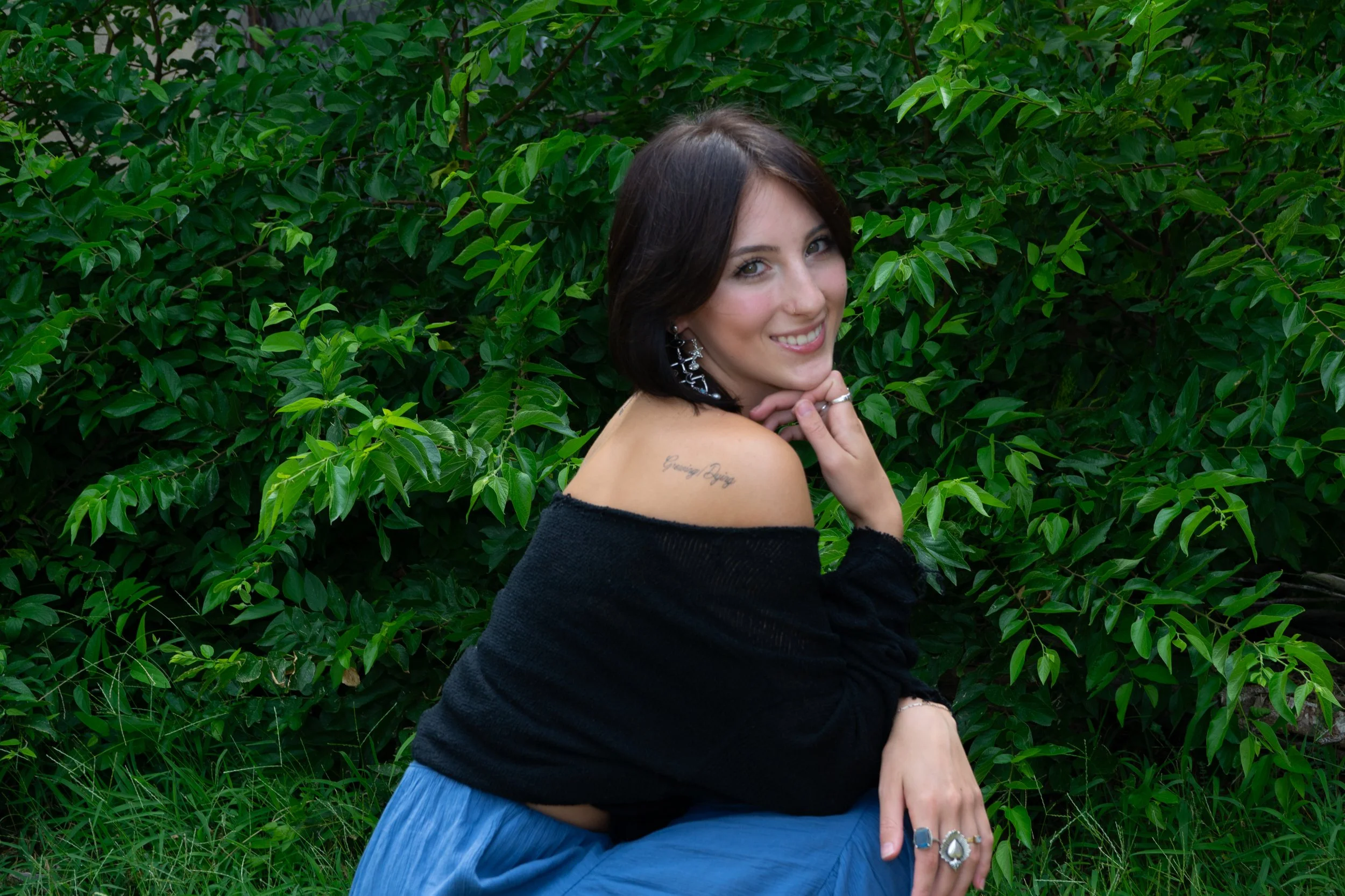 A woman with dark hair and a tattoo on her shoulder, wearing large earrings and rings, is sitting in front of lush green foliage, smiling at the camera.