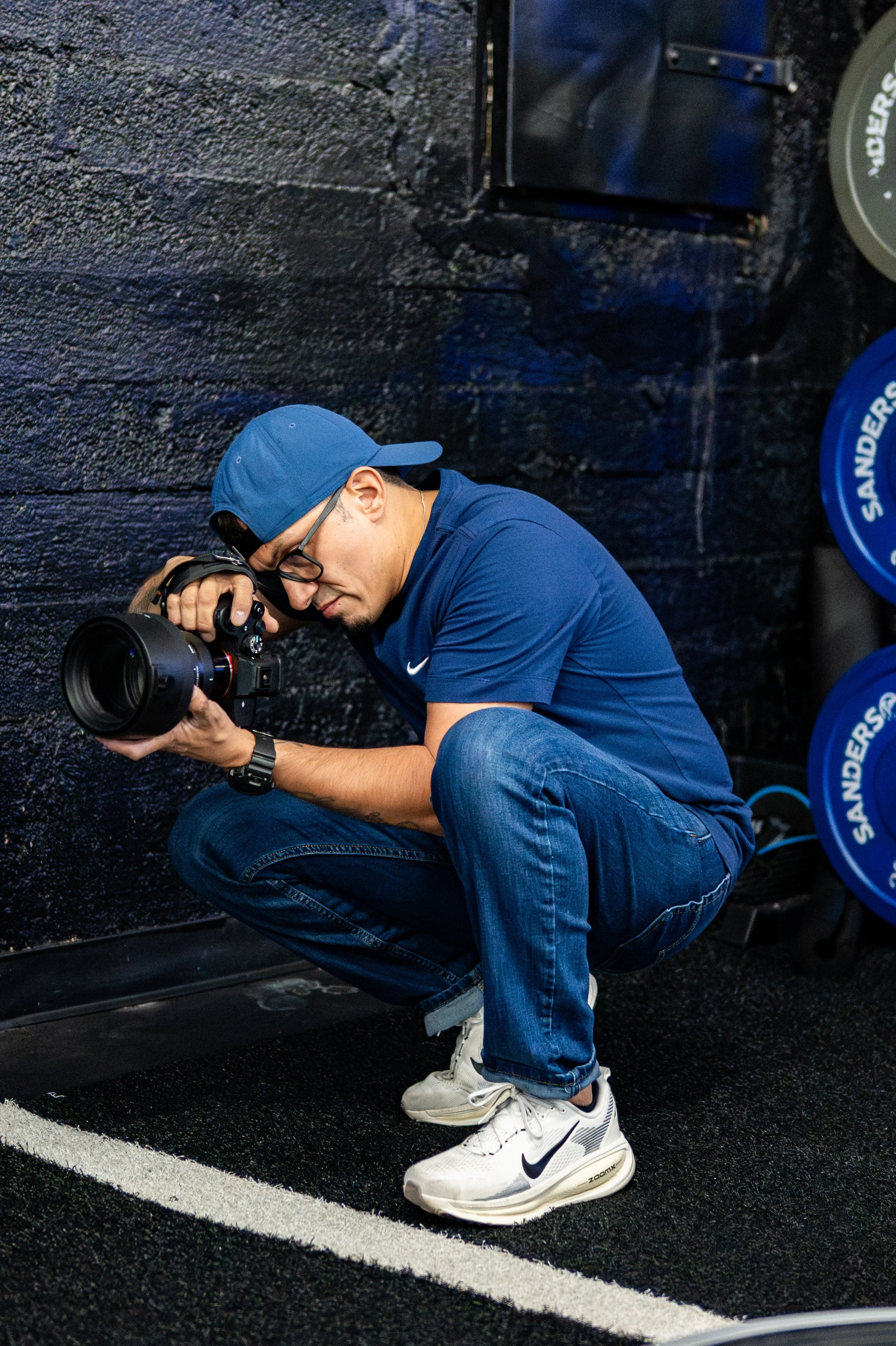 A young man squatting down, holding a camera with a large lens, and taking a photograph against a black brick wall background.