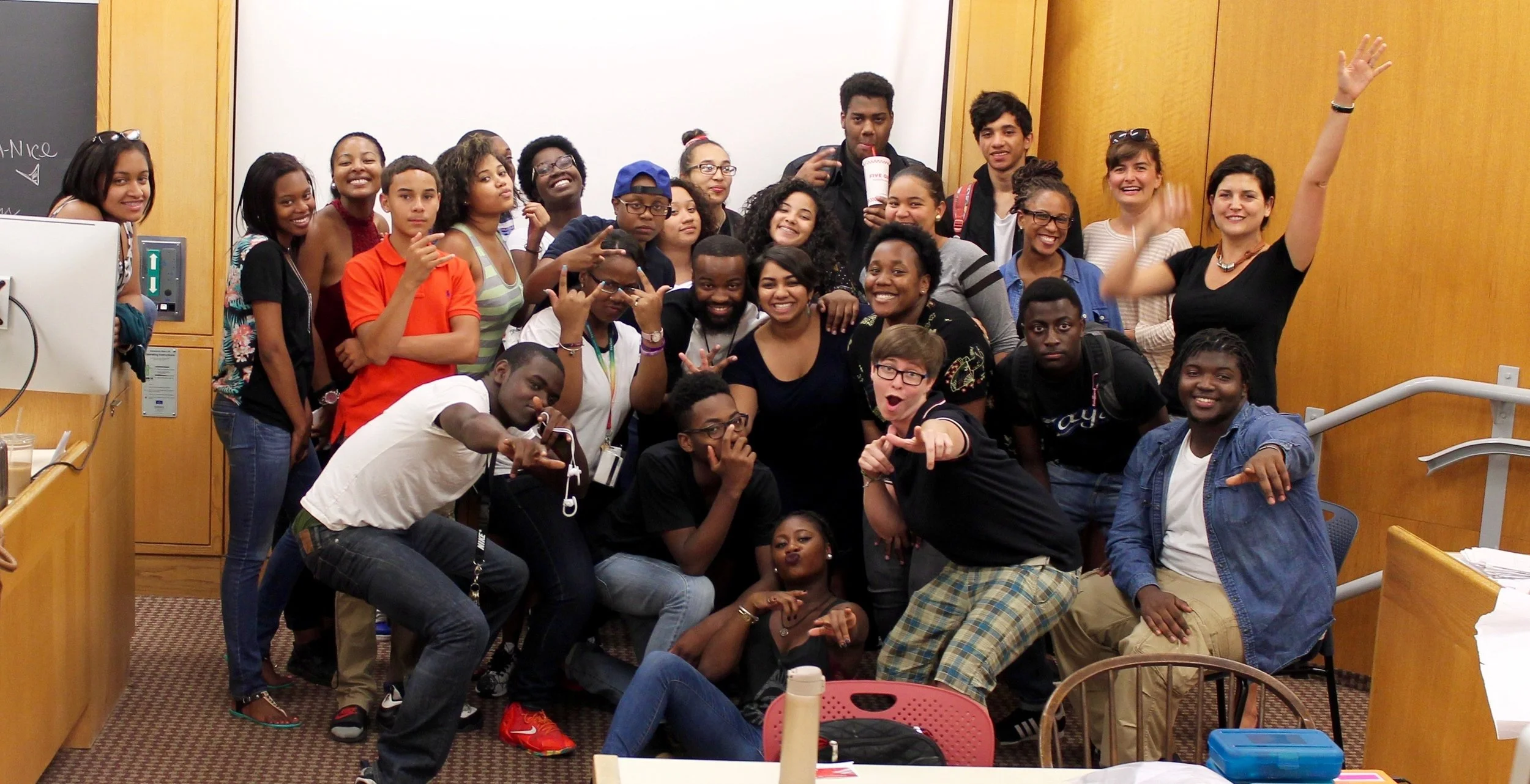 A diverse group of young people and adults posing together in a classroom or lecture hall, smiling, making gestures, and enjoying each other's company.