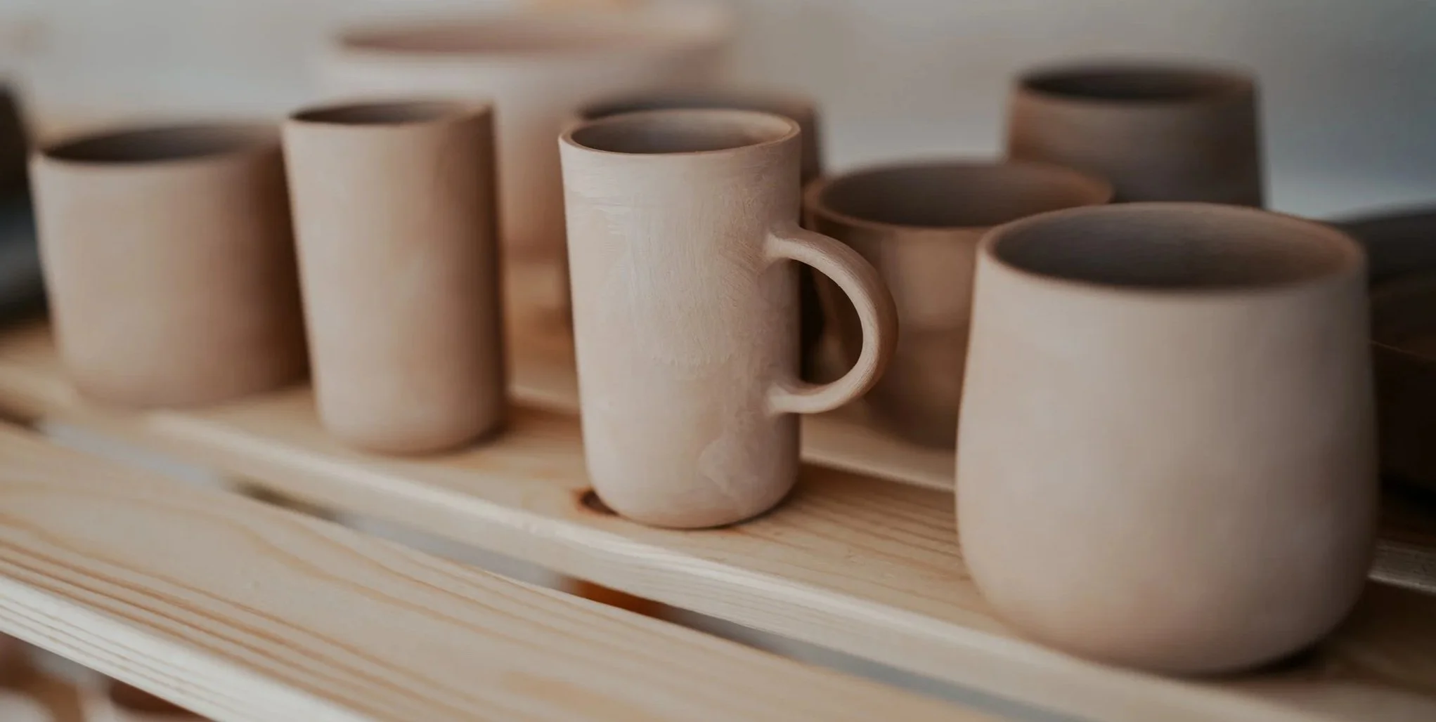 Handmade pottery mugs drying on some wooden sheves, waiting to be fired in the kiln.