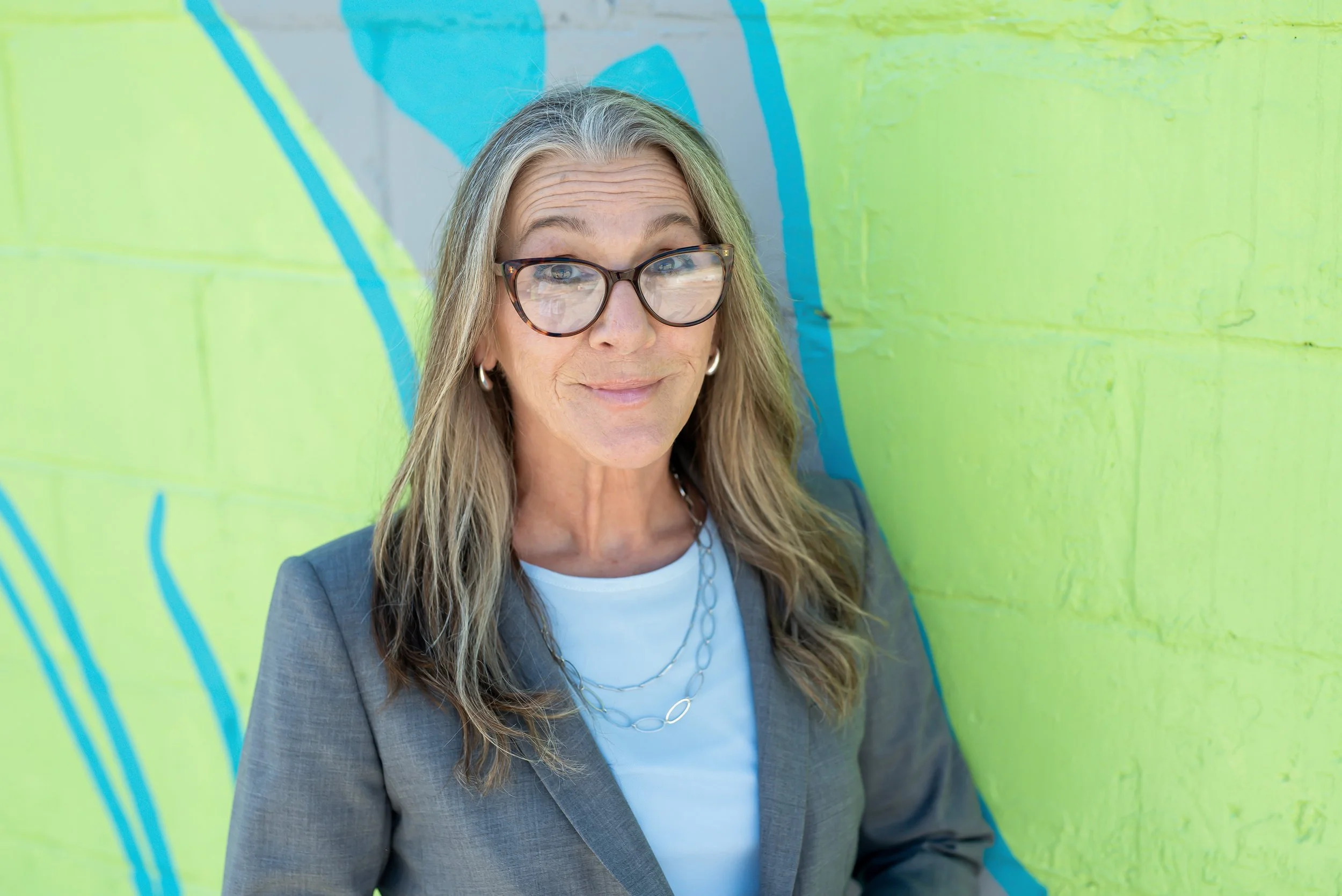 A middle-aged woman with gray hair and glasses, smiling at the camera against a gray background, wearing a black blazer and light blouse.