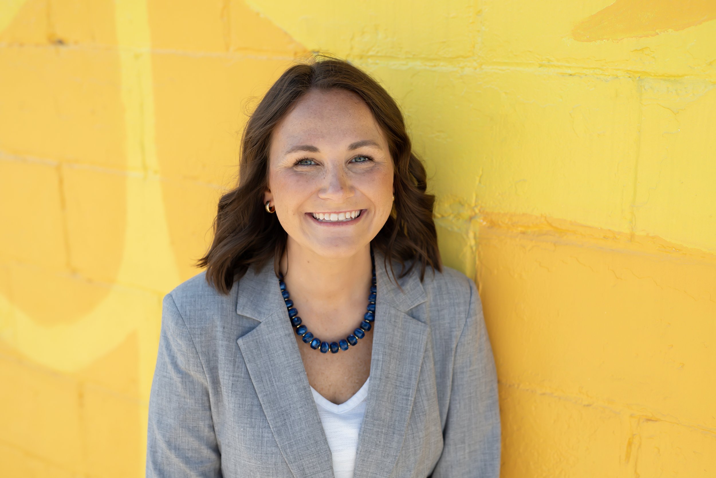 A woman with long brown hair, blue eyes, and a friendly smile, wearing a light gray blazer, a white shirt, and a round badge with a red, blue, and yellow logo. The background is a dark gray gradient.