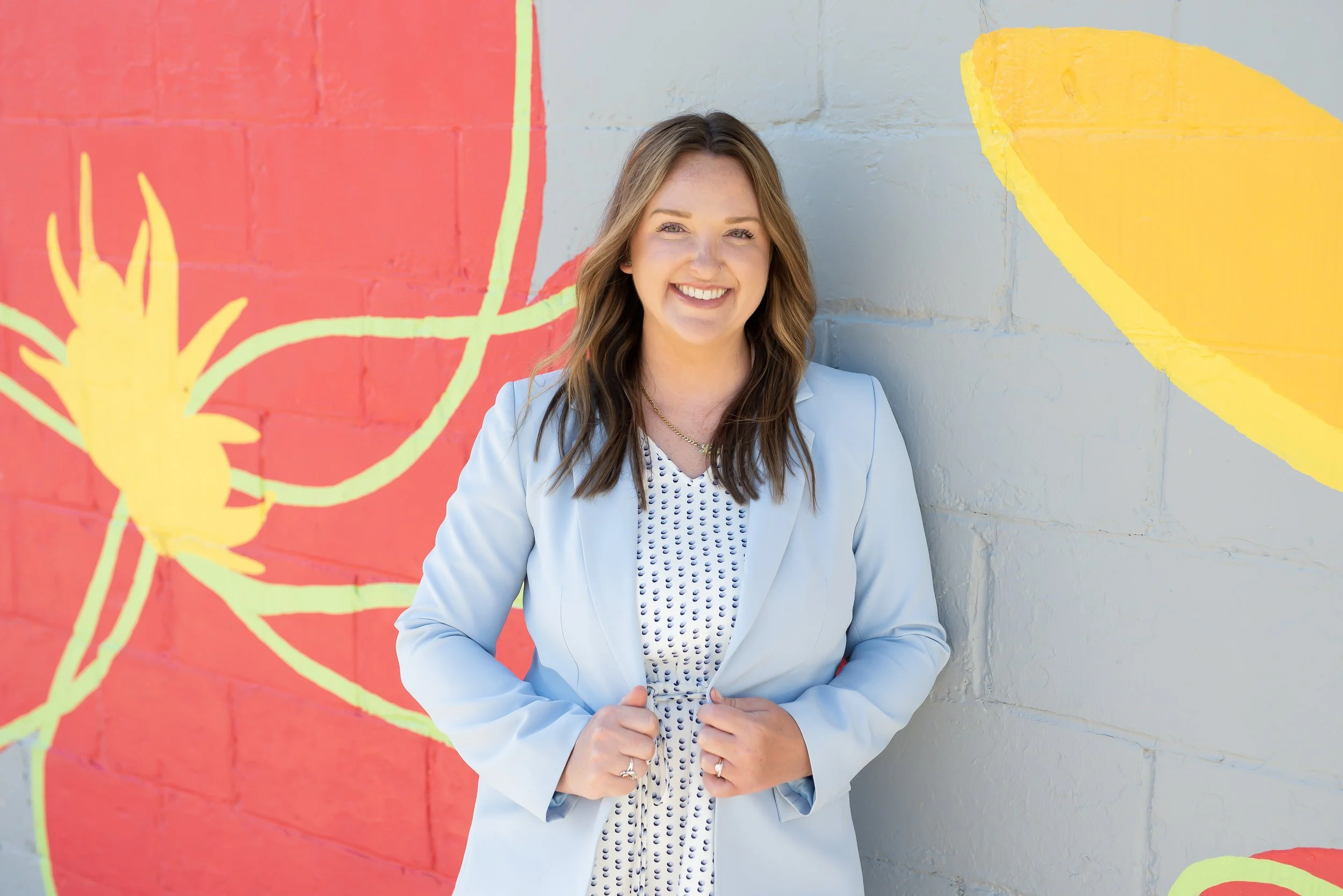 A young woman with long brown hair, blue eyes, and light skin, smiling, wearing a light blue blazer over a white blouse with floral embroidery, and a colorful pin on her blazer, against a gray background.