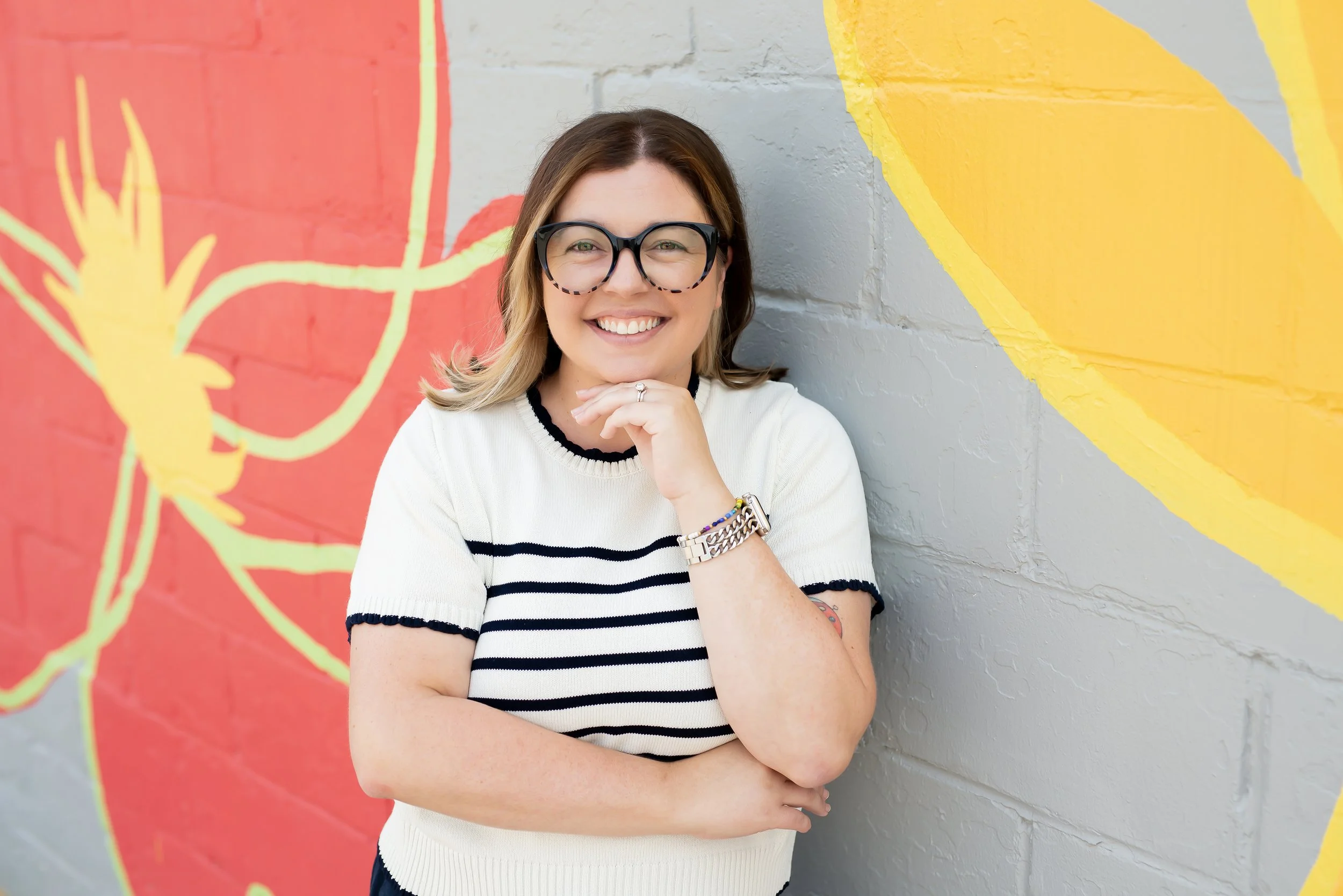 Portrait of a woman with wavy dark hair, wearing blue glasses, smiling, in a light beige blouse with polka dots, against a gray textured backdrop.