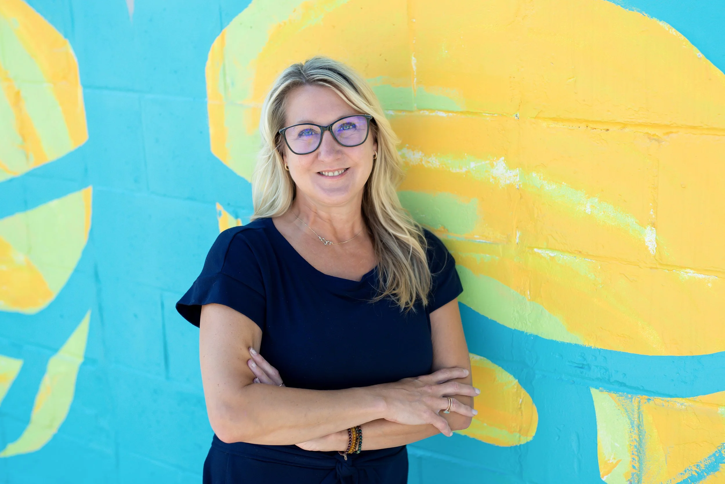 A woman with long blonde hair wearing glasses, smiling, against a colorful background.