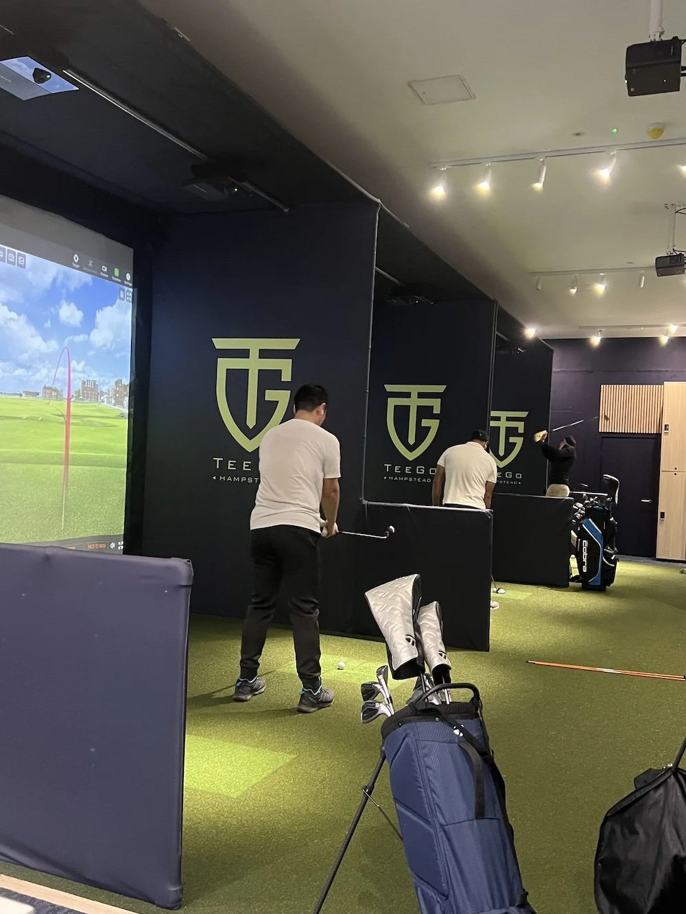 Golfers practising in a TeeGo simulator bay in front of the TeeGo logo wall