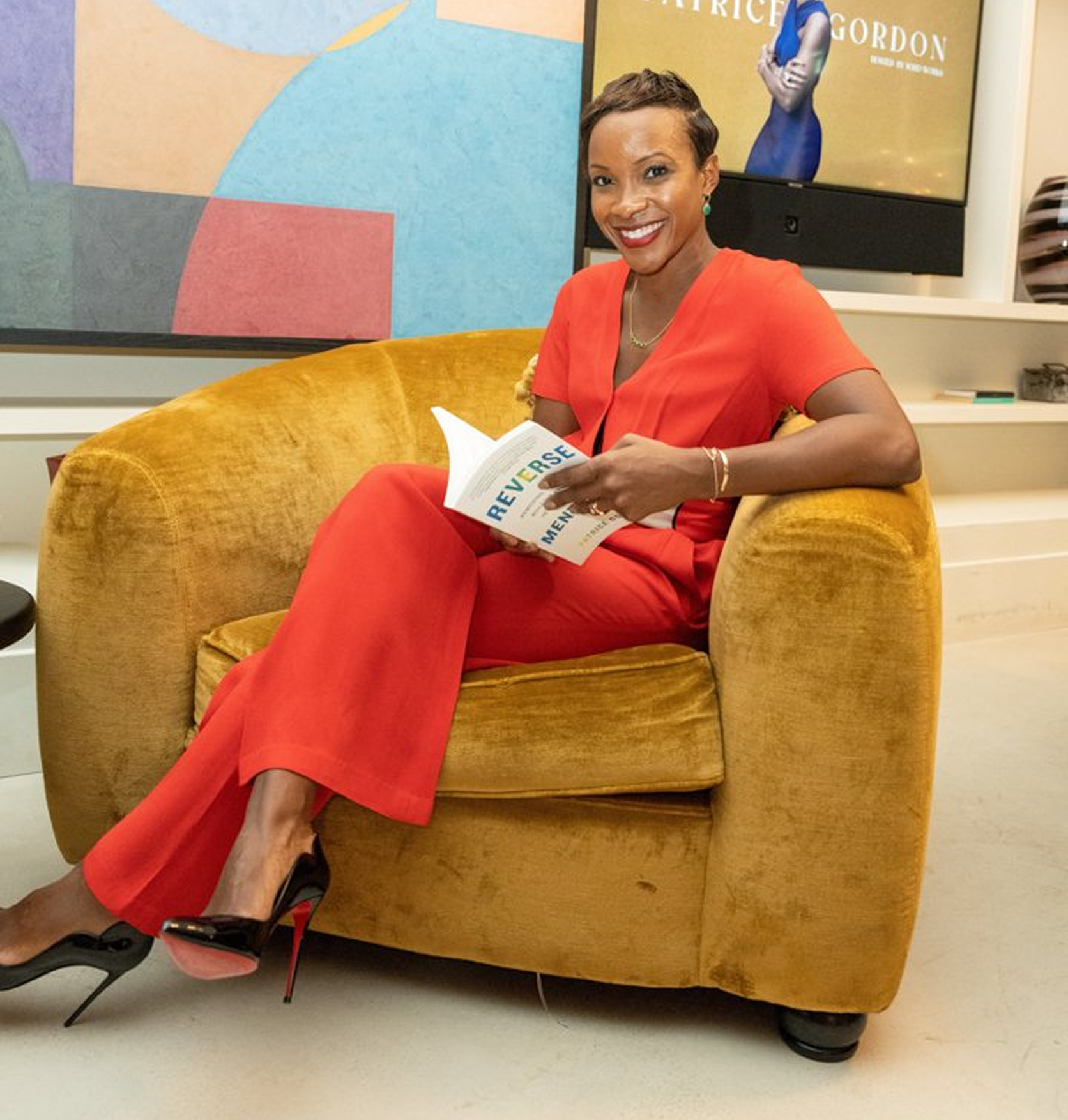 Patrice Gordon reading a book titled "Reverse" in a modern indoor setting with artwork and a television in the background.