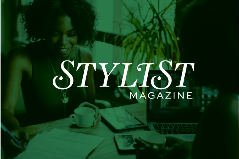 A woman with curly hair smiling at a table with a coffee mug, notes, smartphone, laptop, and a tablet in front of her, with the text 'Stylist Magazine' overlayed.