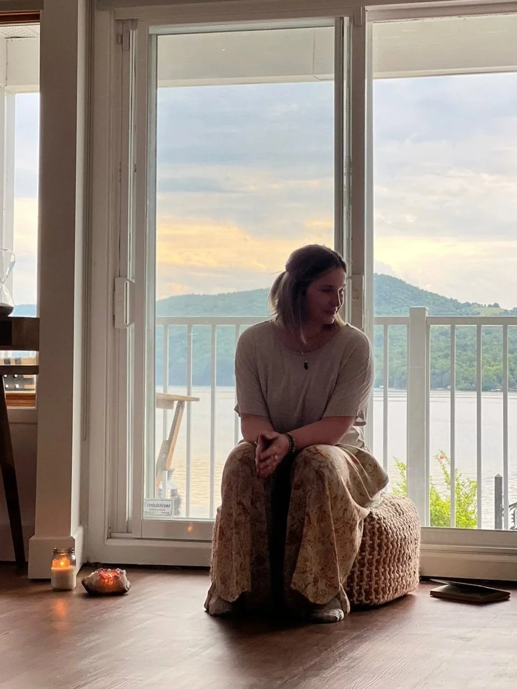 A woman sitting on a textured cushion near a sliding glass door with a view of a lake, mountains, and a cloudy sky in the background.