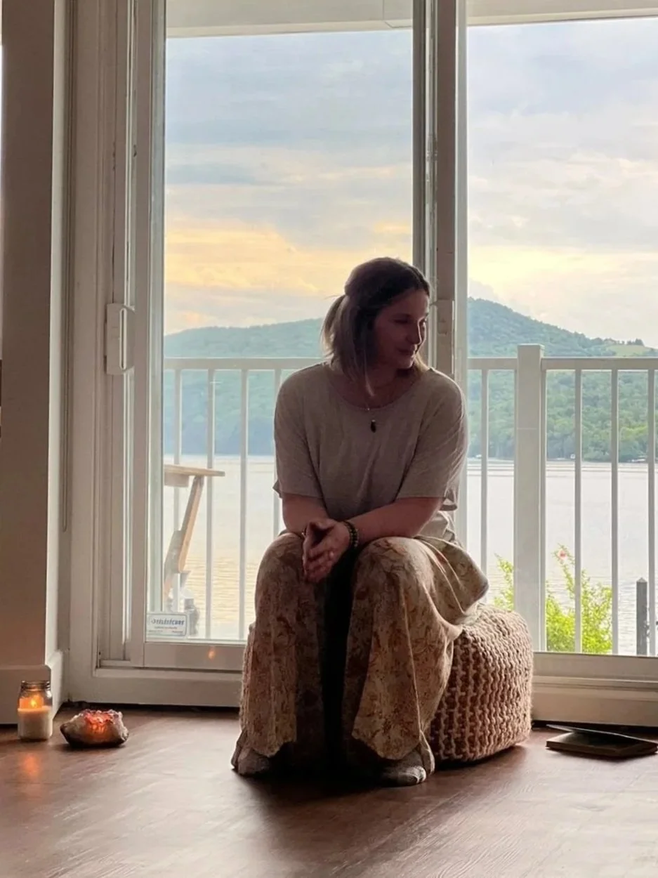 A woman sitting on a woven ottoman near a glass door overlooking a river and mountains during sunset.