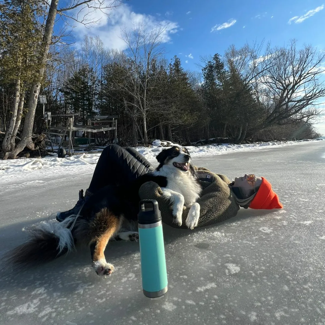 A person lying on their back on a frozen lake with a Border Collie dog resting its head and paw on the person's chest. The person is wearing a red hat and sunglasses with a blanket or coat. A water bottle is in the foreground. In the background, there are trees and some outdoor furniture under a partly cloudy sky.