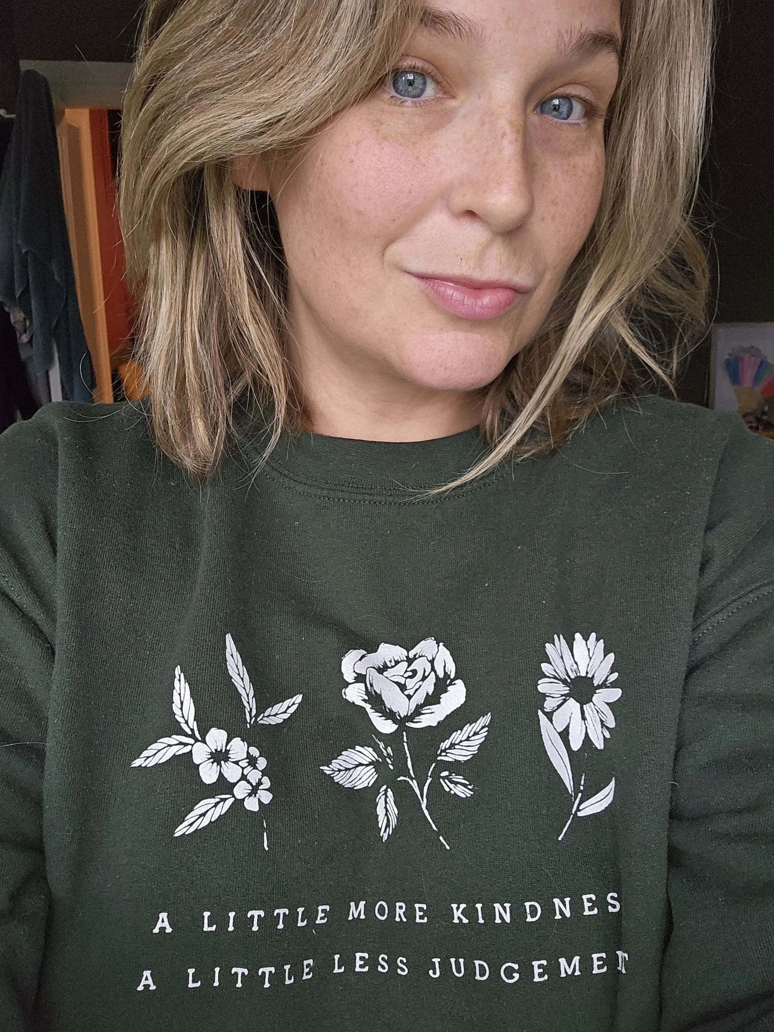 Close-up of a woman with blonde hair and blue eyes, wearing a black sweatshirt with white floral illustrations and motivational text.