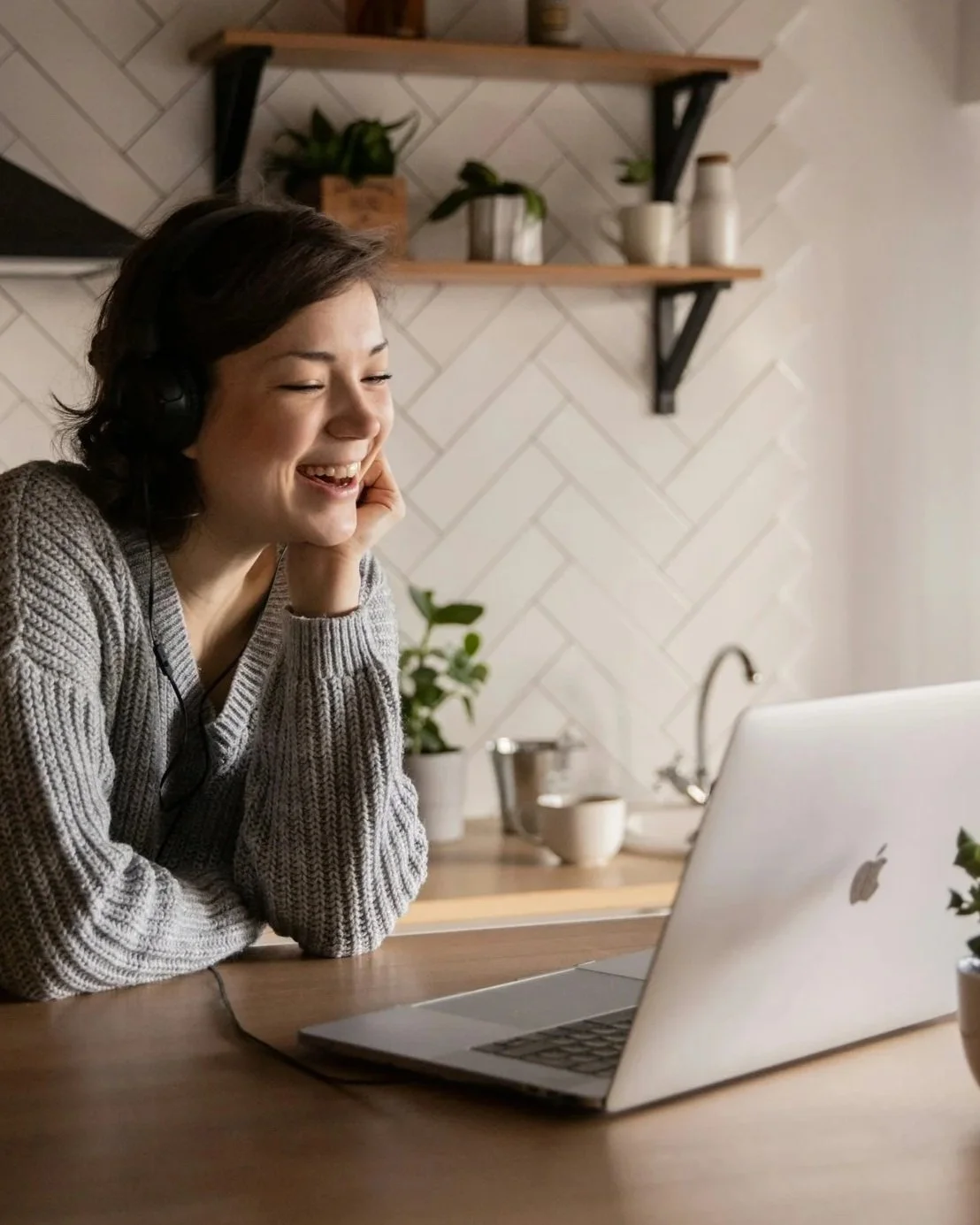 Woman smiling while wearing headphones, sitting at a wooden table with a laptop in a kitchen.