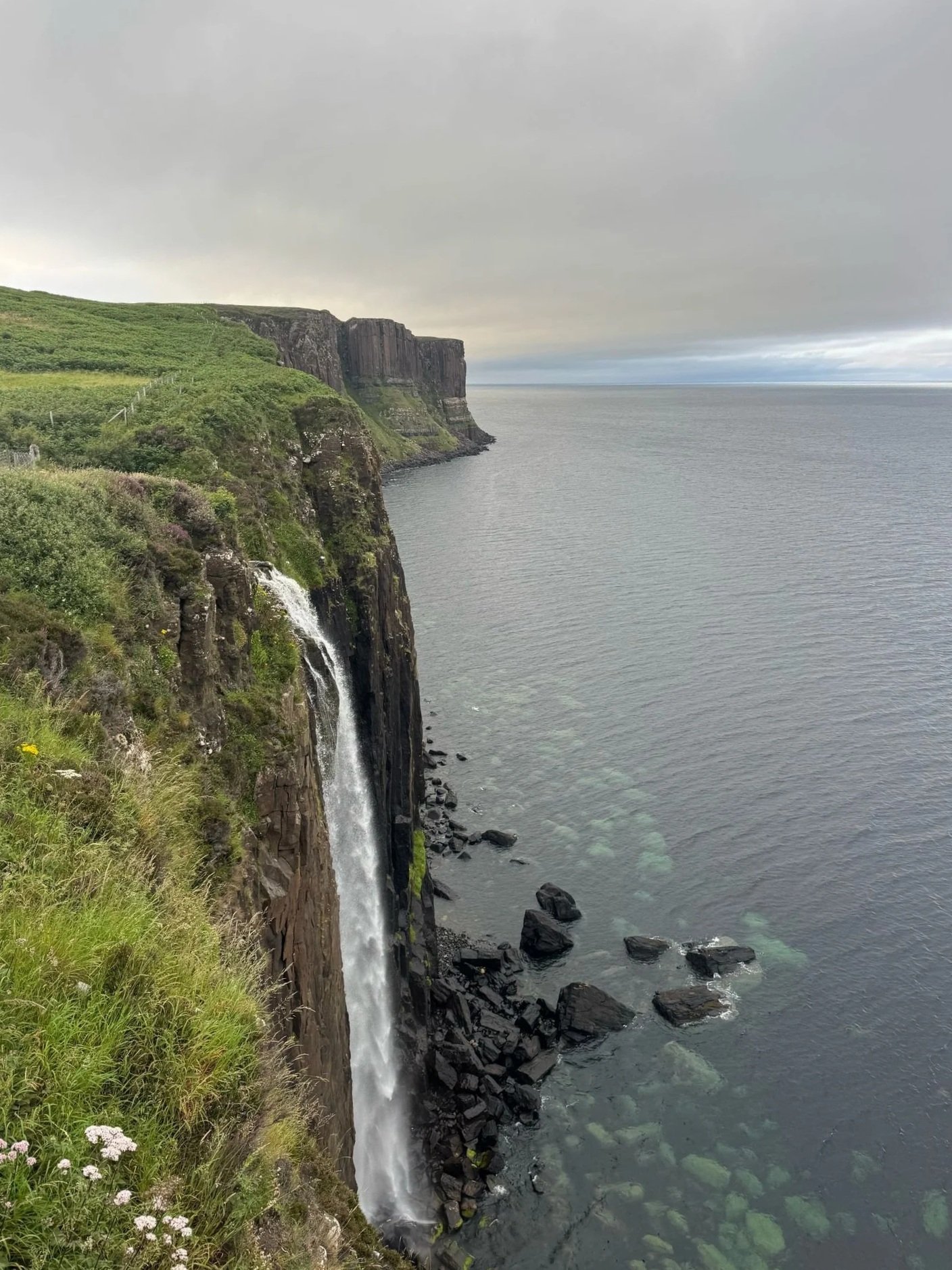 A coastal scene with steep green cliffs, a waterfall flowing down the cliffside, and the ocean below under an overcast sky.