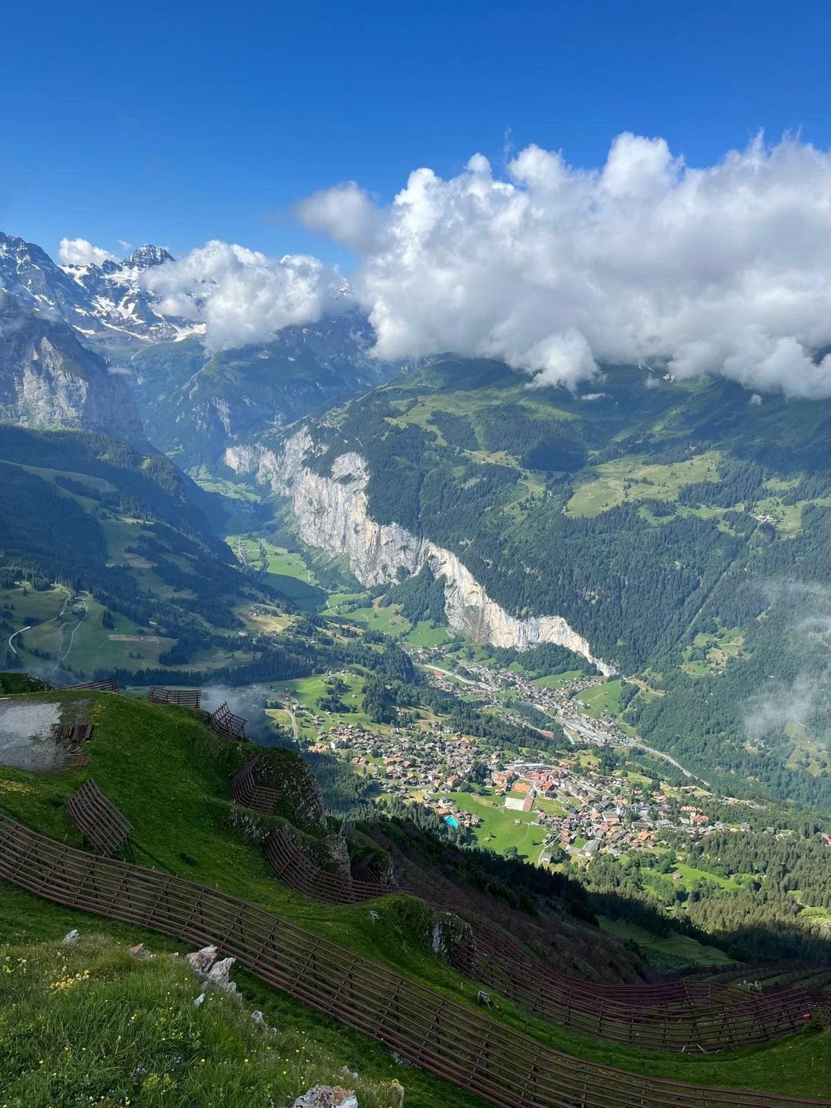 Mountain valley with snow-capped peaks, green hills, and a small town, under a partly cloudy blue sky.
