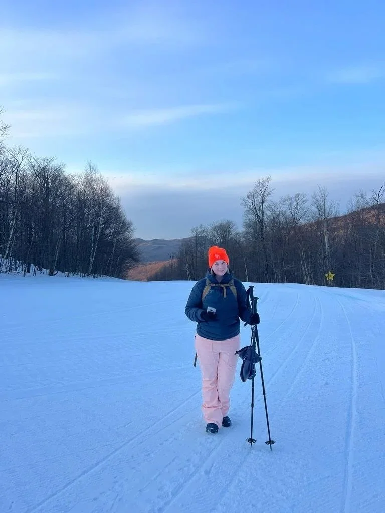 Woman in a bright orange hat, navy jacket, pink pants, and black gloves walking on a snowy trail with trekking poles. She is holding a phone and carrying a backpack. Bare trees and hills are in the background under a partly cloudy sky.