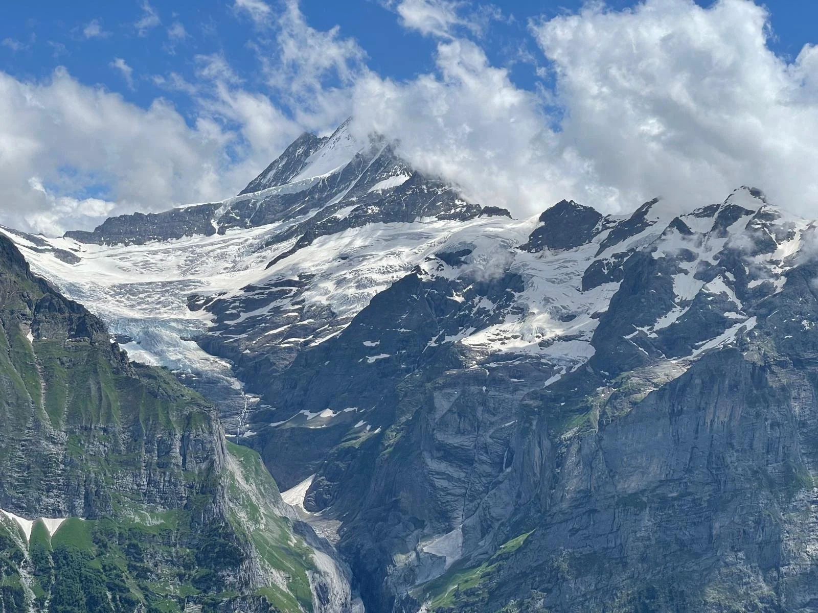 Snow-capped mountain with clouds around the peak and green slopes at the base.