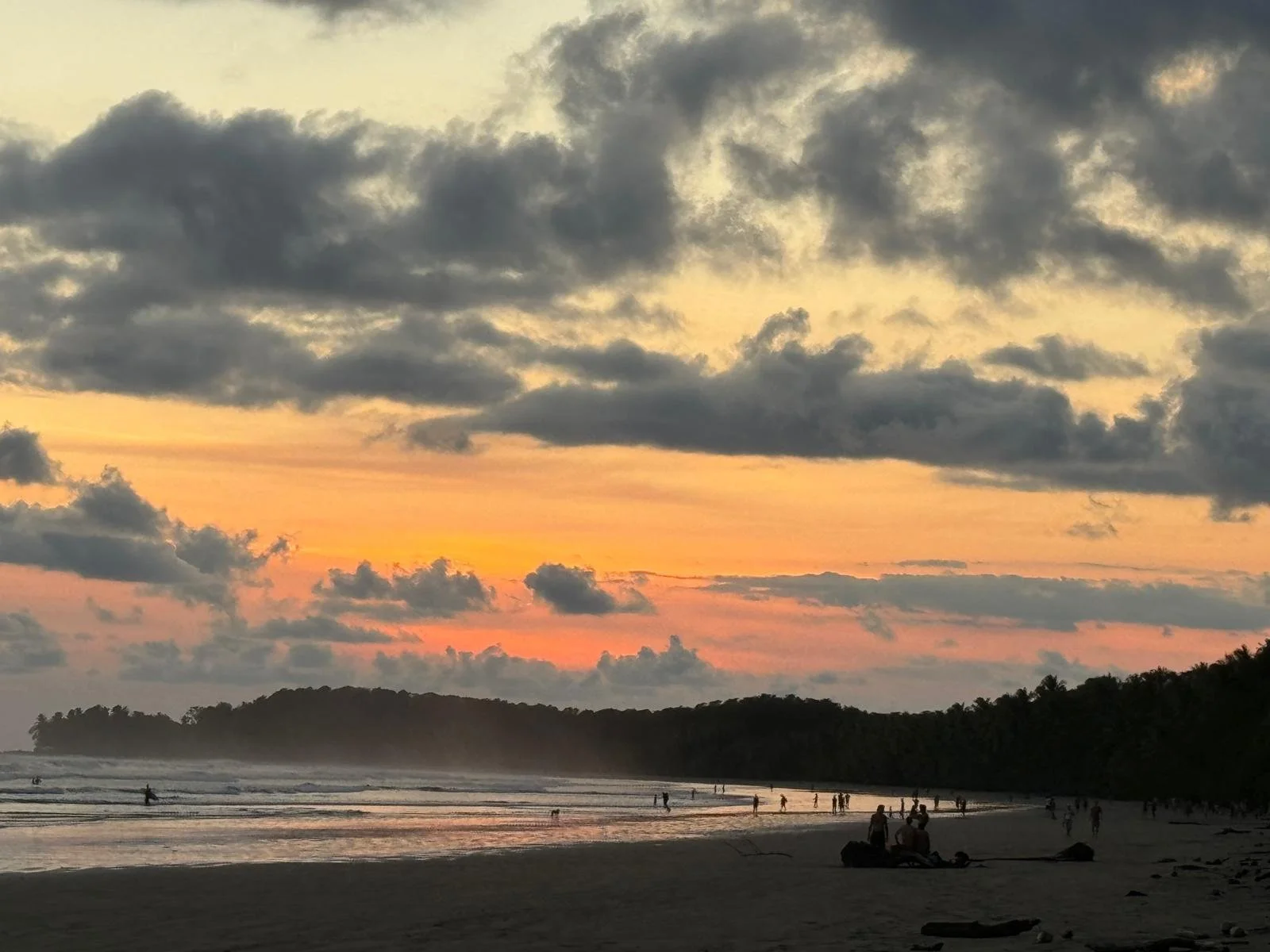 Sunset over a beach with clouds in the sky, people walking and sitting on the sand, and hills in the background.