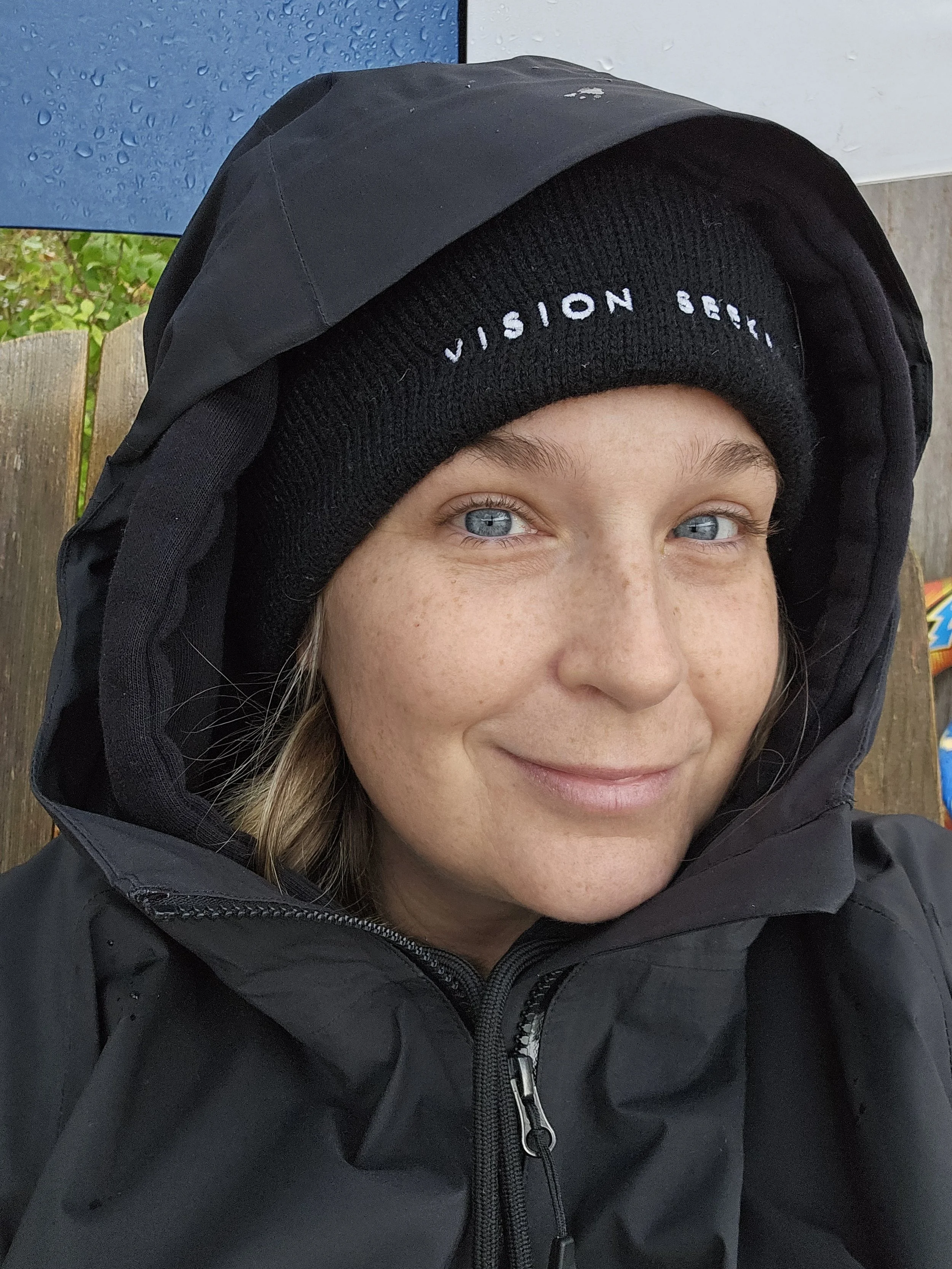 Close-up of a woman with light skin and blue eyes wearing a black beanie with white embroidered text and a black jacket, smiling slightly, with a background featuring blue and green elements.
