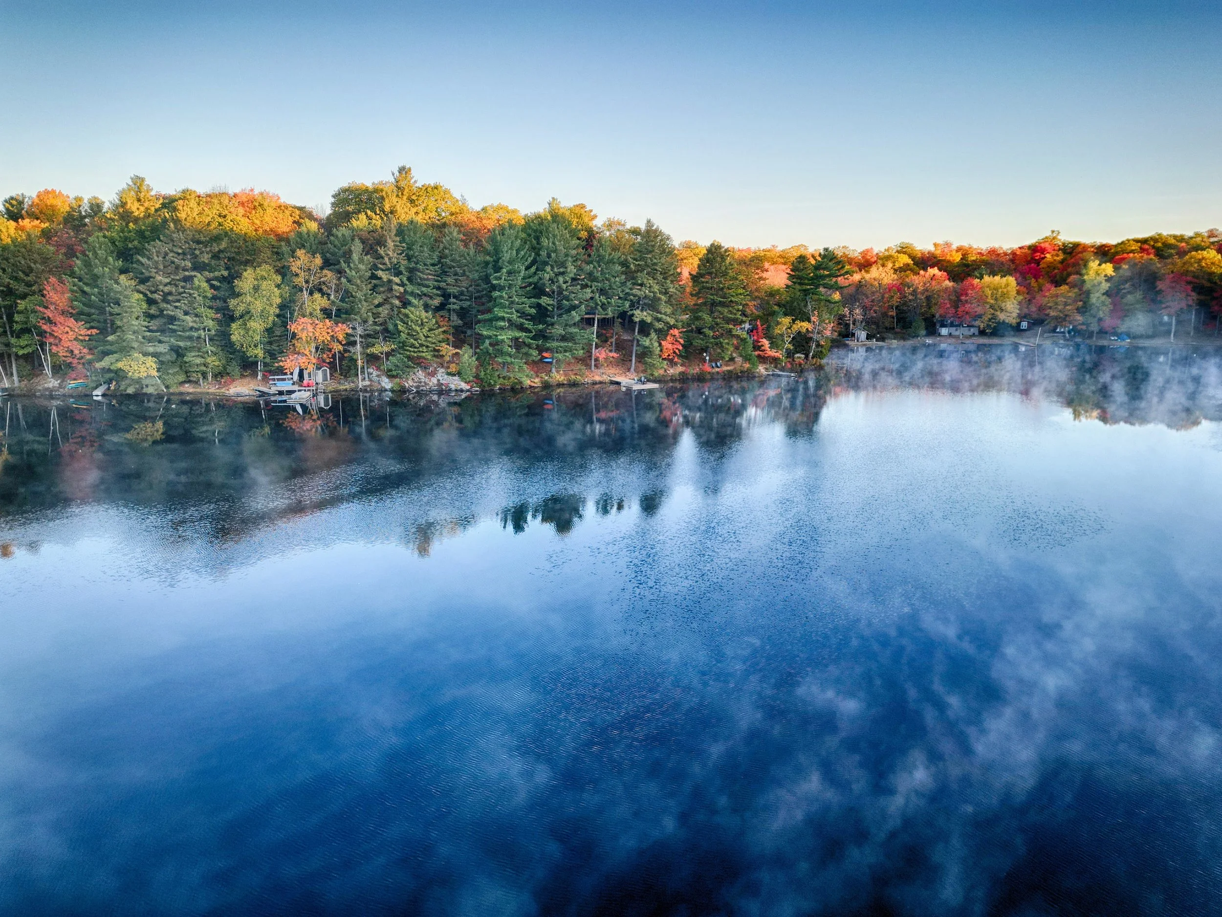 A calm lake reflecting the colorful trees along the shoreline with fog rising from the water in the early morning.
