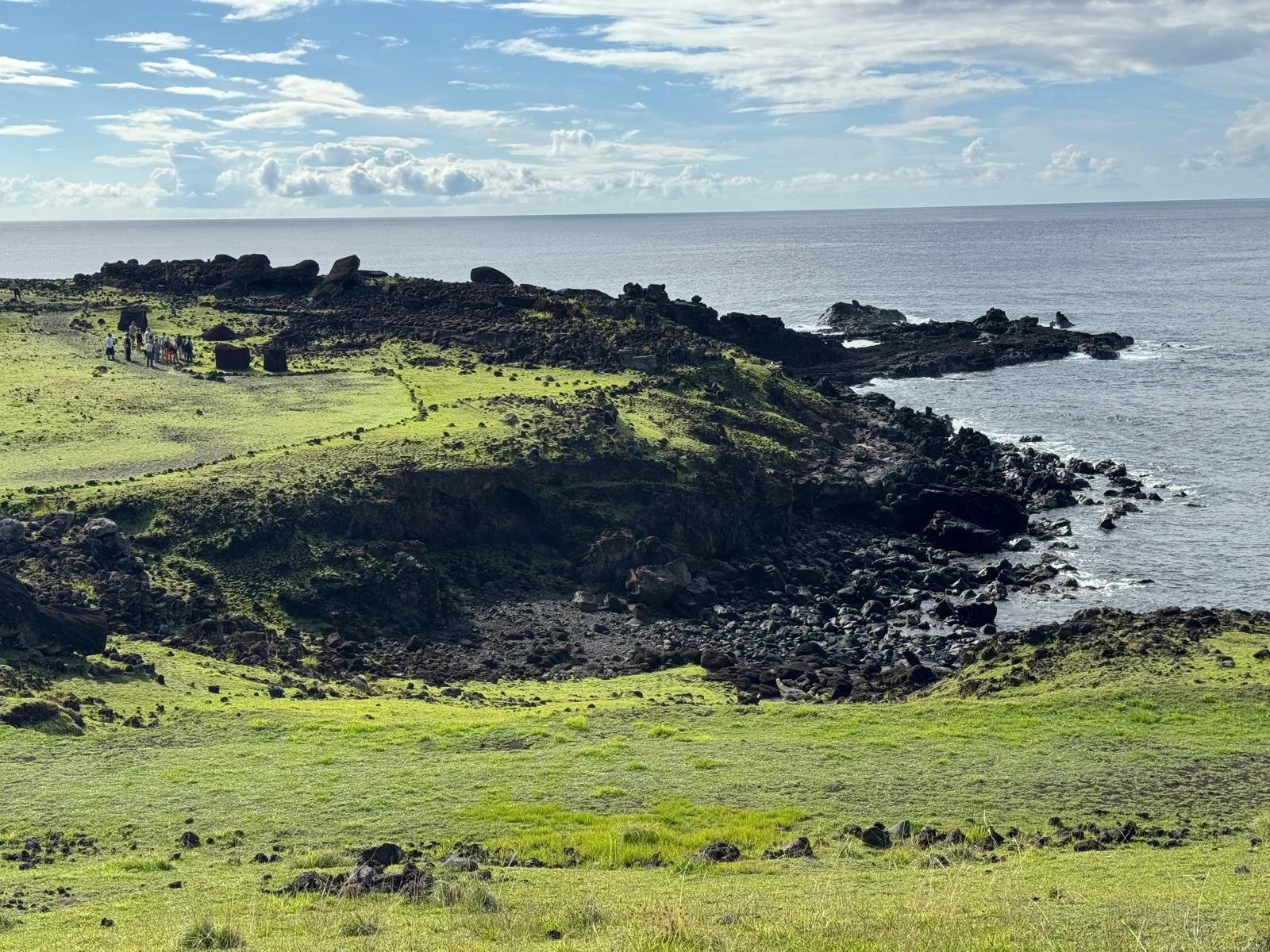 A coastal landscape with grassy green hills, rocky shoreline, and ocean view under partly cloudy sky, with a group of people near the ruins of an ancient structure.