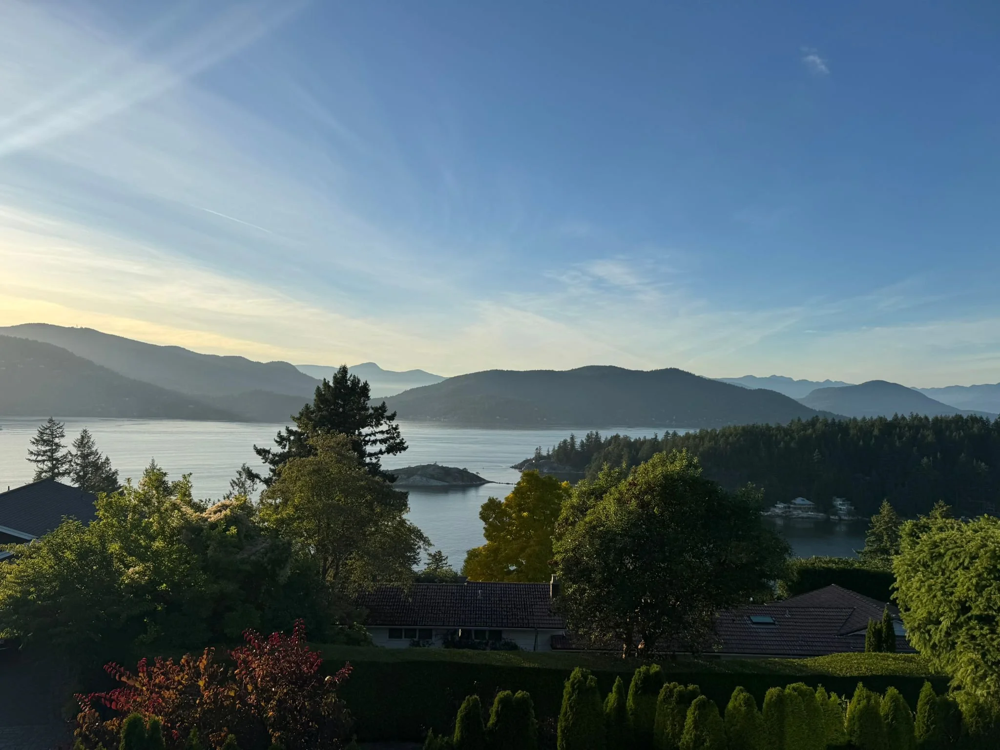 View of a large body of water with surrounding green trees, mountains in the distance, and a partly cloudy sky at sunset.