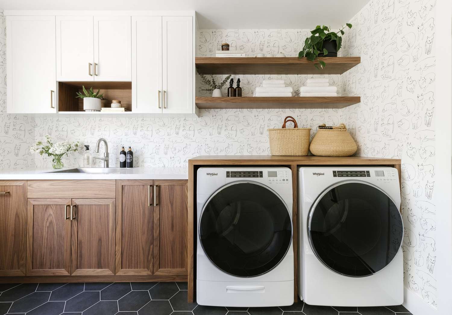 A laundry room with a white countertop and wooden cabinets, open shelves with decor, and two front-loading washing machines. Wall has cat doodle wallpaper, and there are baskets on top of the machines.