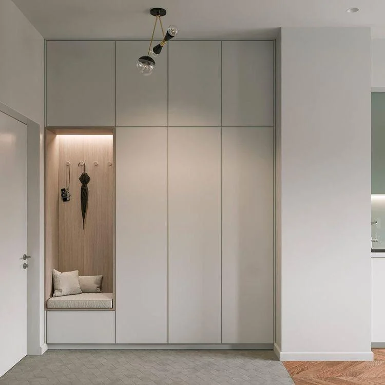 Modern entryway with a built-in nook featuring a cushion, pillows, hooks for hanging items, and a wooden panel background, next to a white wall and a cabinet.